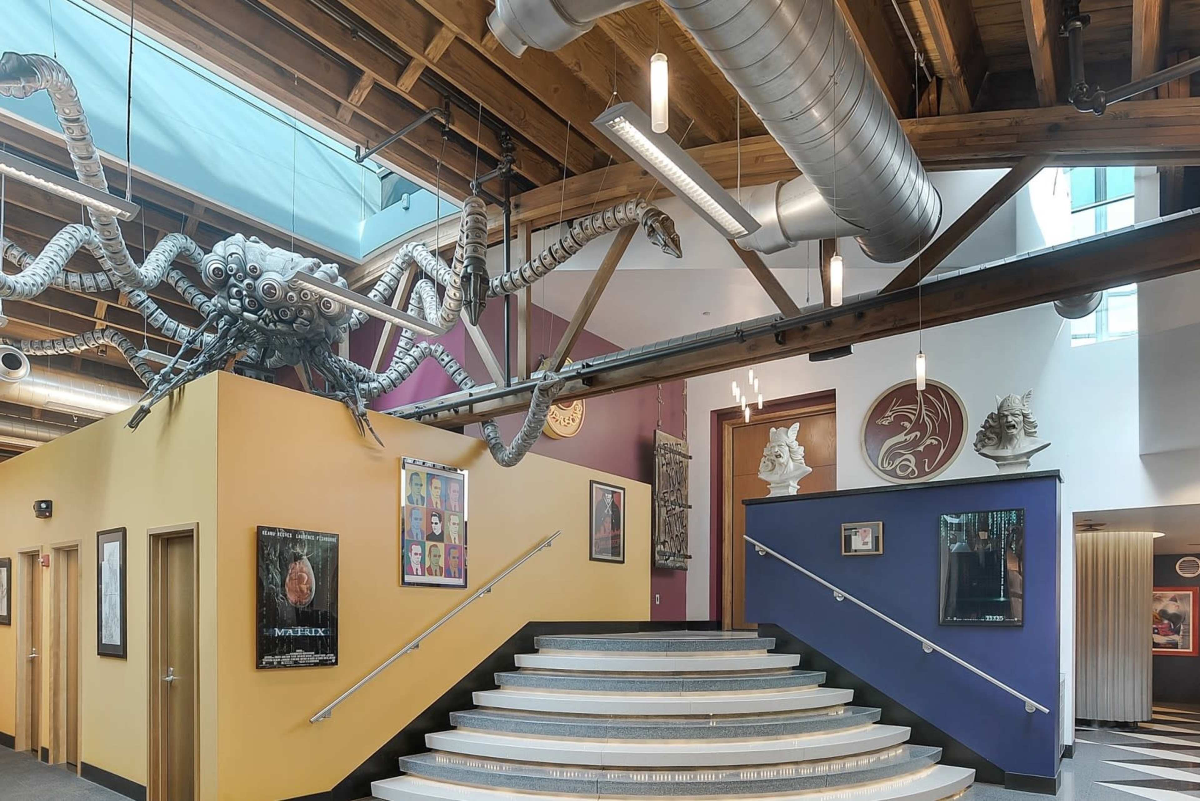 A spacious interior with a staircase leading up, framed by colorful walls and industrial pipes above, featuring a large metallic spider sculpture suspended from the ceiling.