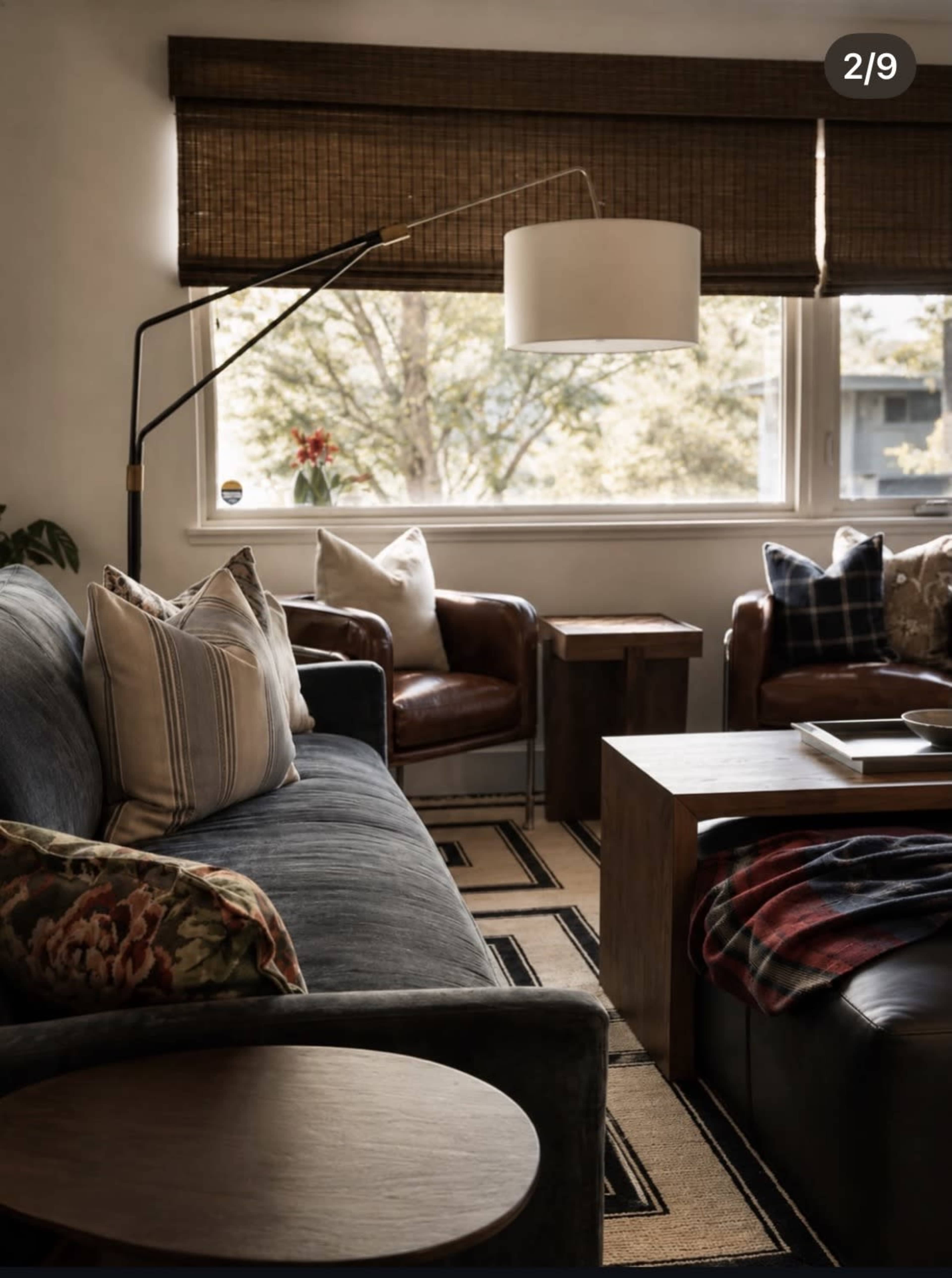 The image shows a cozy living room with a dark sofa, a chair, and a wooden side table, illuminated by natural light from a large window covered with bamboo shades.