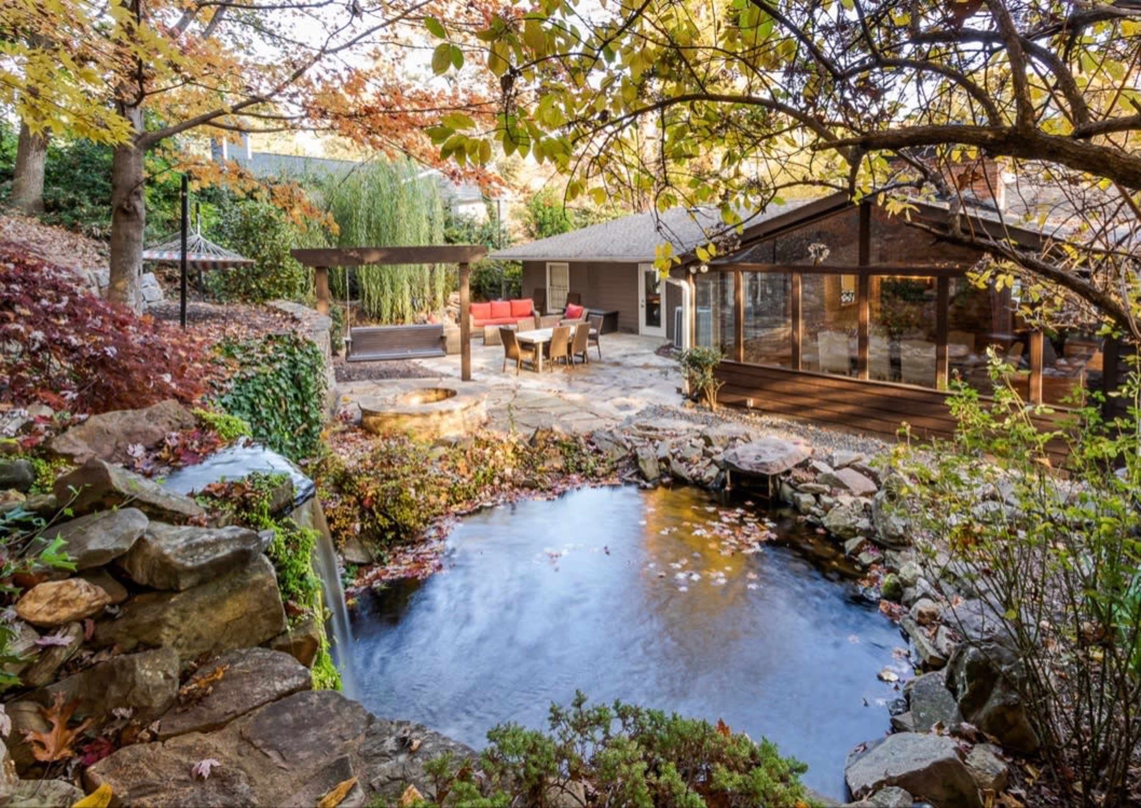 The image shows a landscaped backyard with a pond surrounded by rocks and autumn foliage, featuring a wooden deck and seated area.