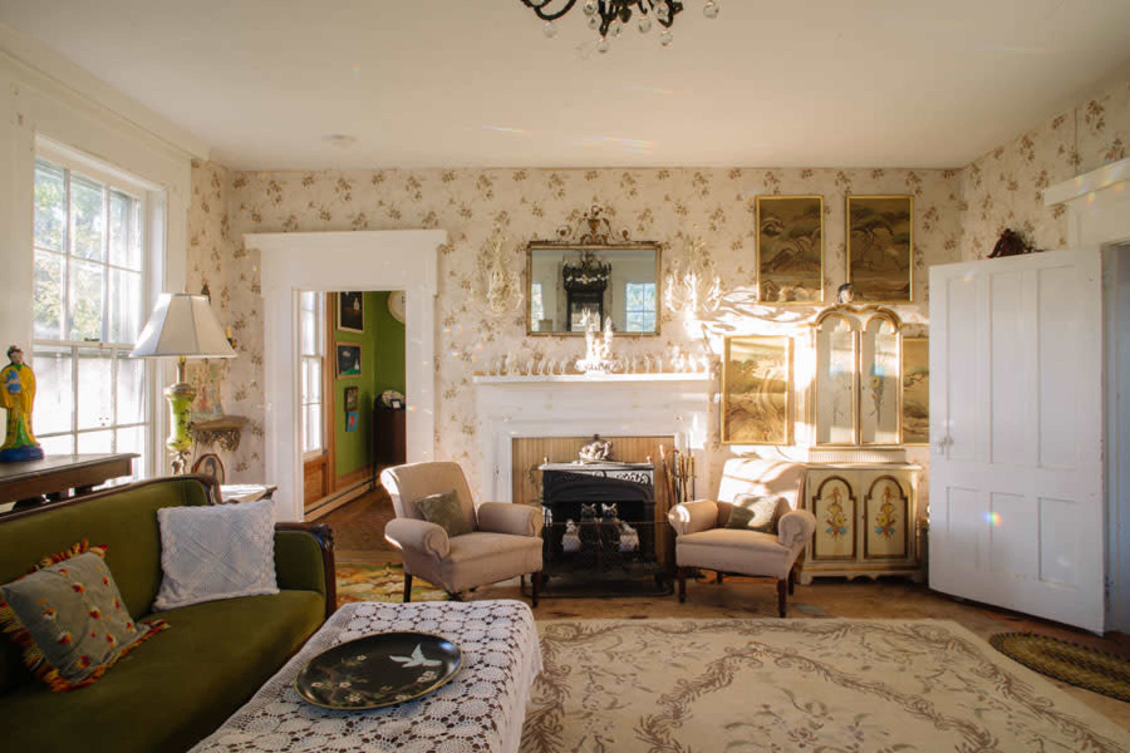 The room features a vintage sofa, a decorative rug, a fireplace with a mantle, and a large cabinet illuminated by sunlight.