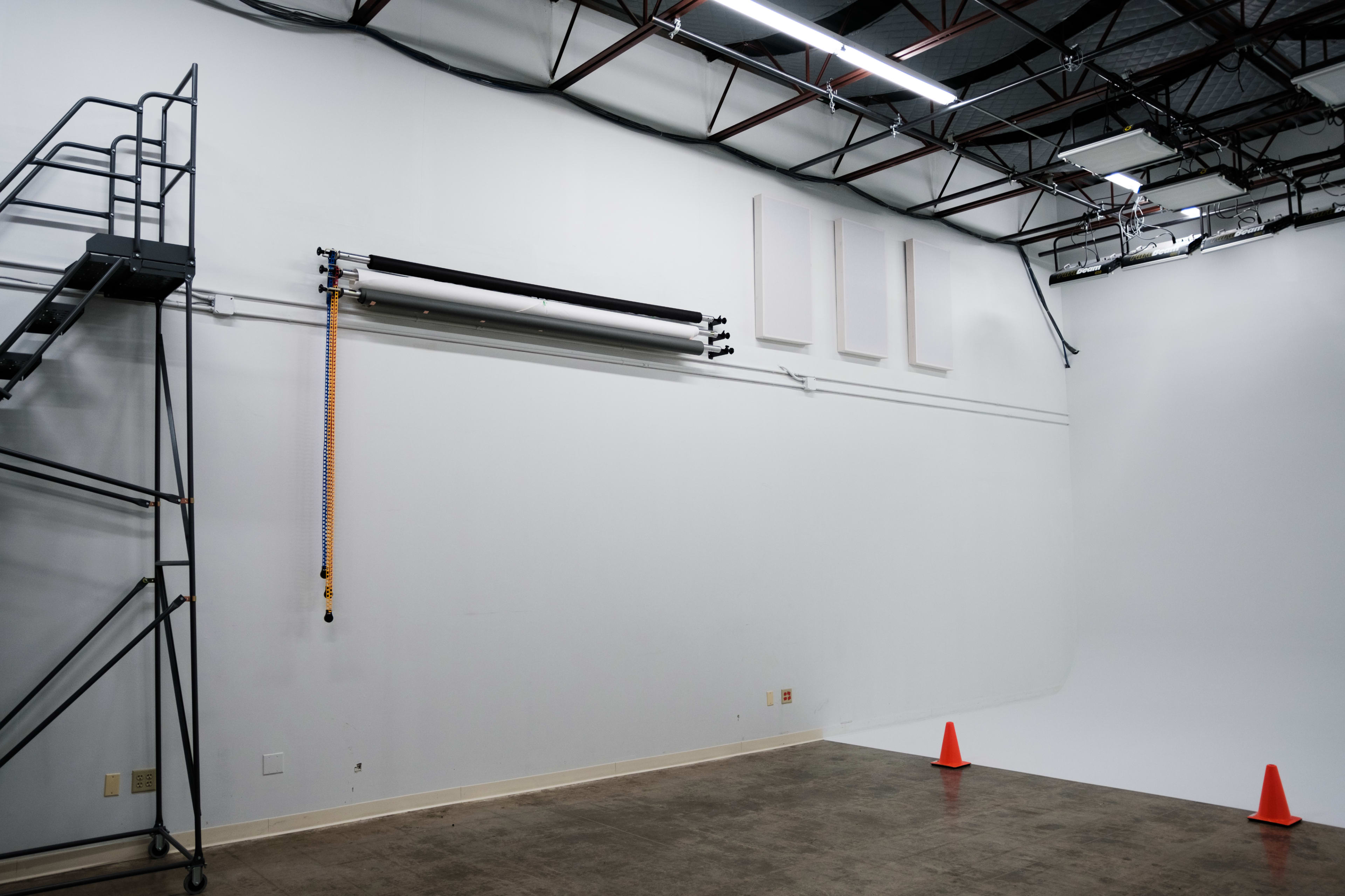 An empty photography studio with a white backdrop, a ladder against the wall, and orange cones on the floor.