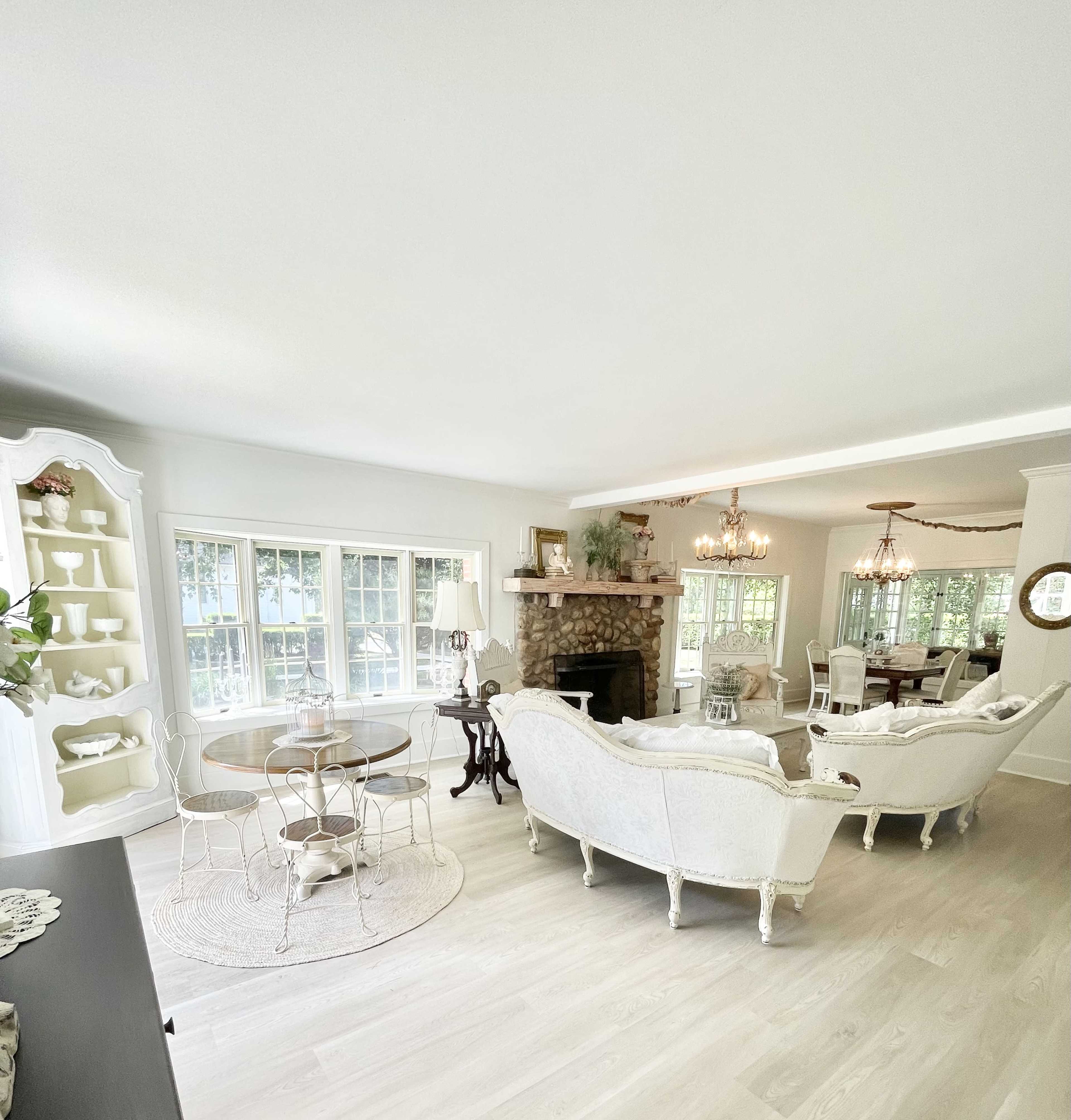 A bright living room with natural light, featuring a stone fireplace, white furniture, and a round dining table set near a large window.