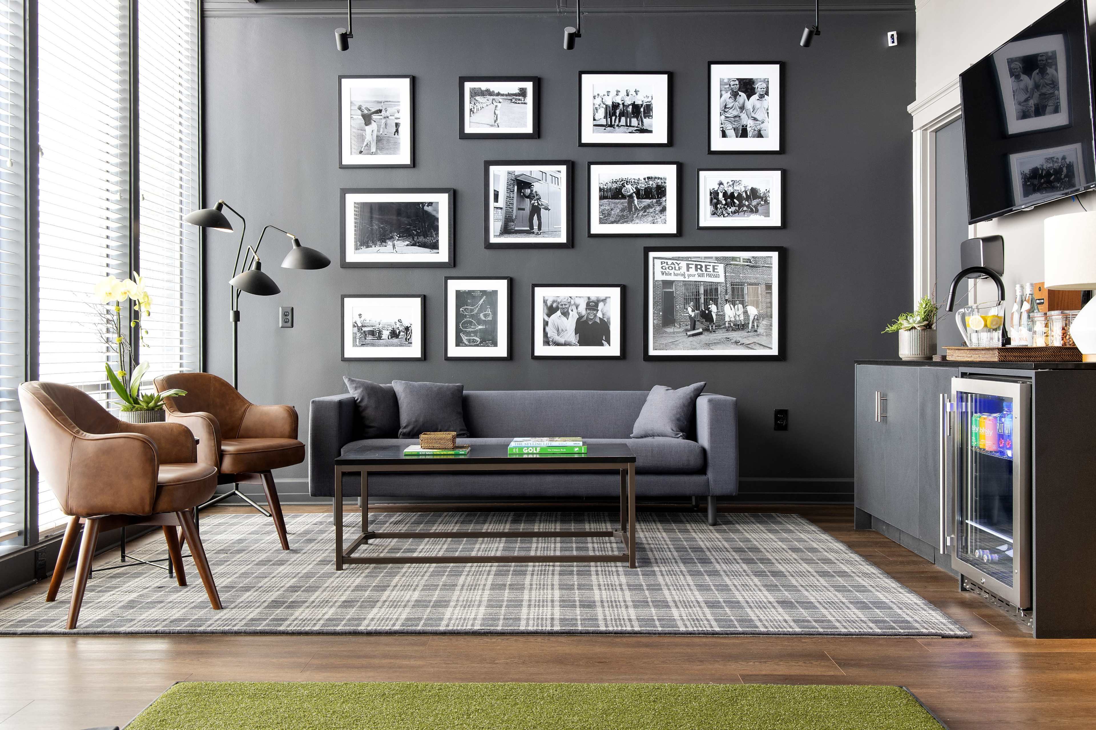 A modern living room with a dark gray wall featuring framed black-and-white photos, a gray sofa, two brown chairs, a coffee table, and a mini fridge.