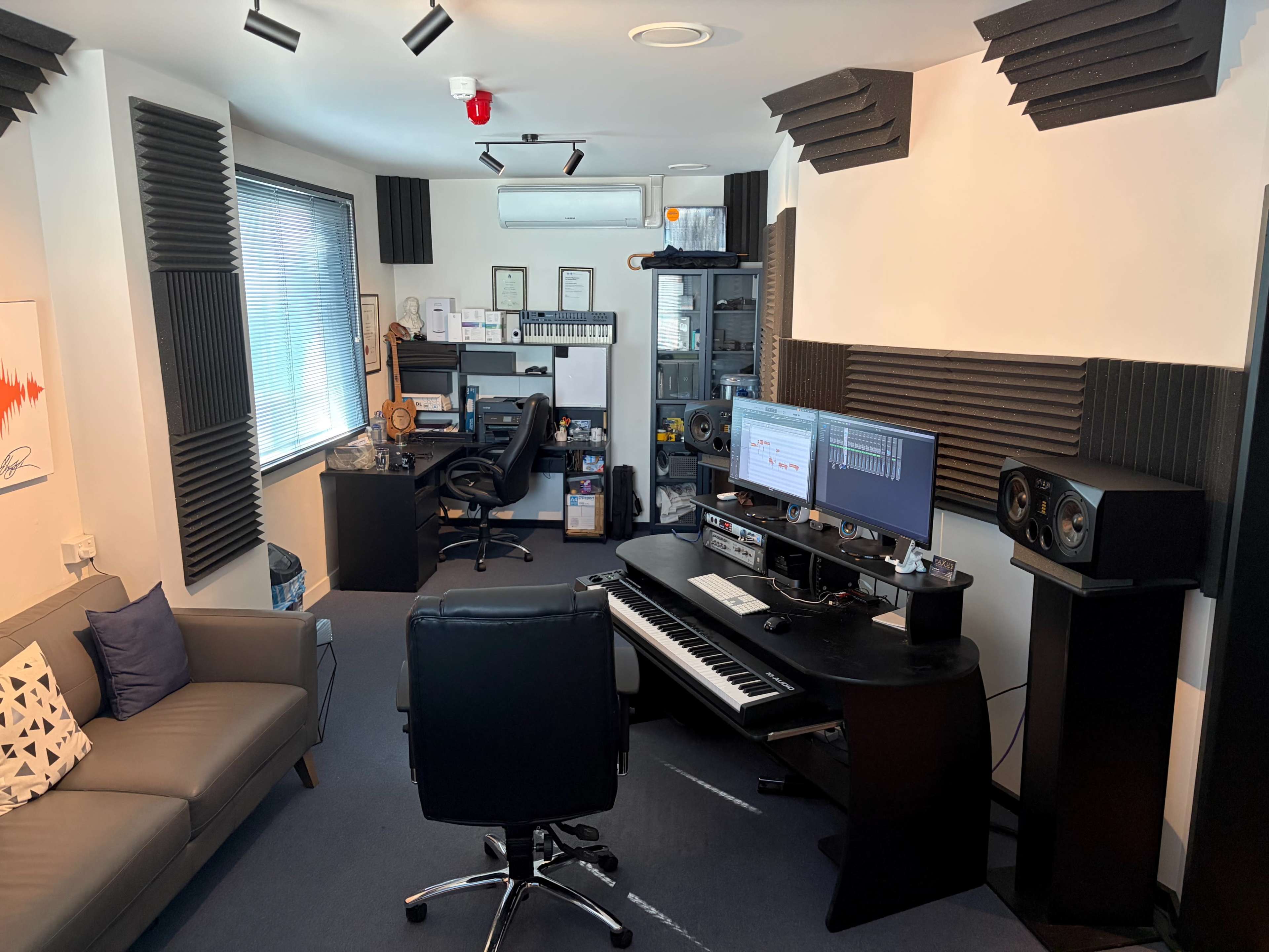 The image shows a music production studio with acoustic foam panels on the walls, a keyboard, two monitors, and a sitting area with a couch.
