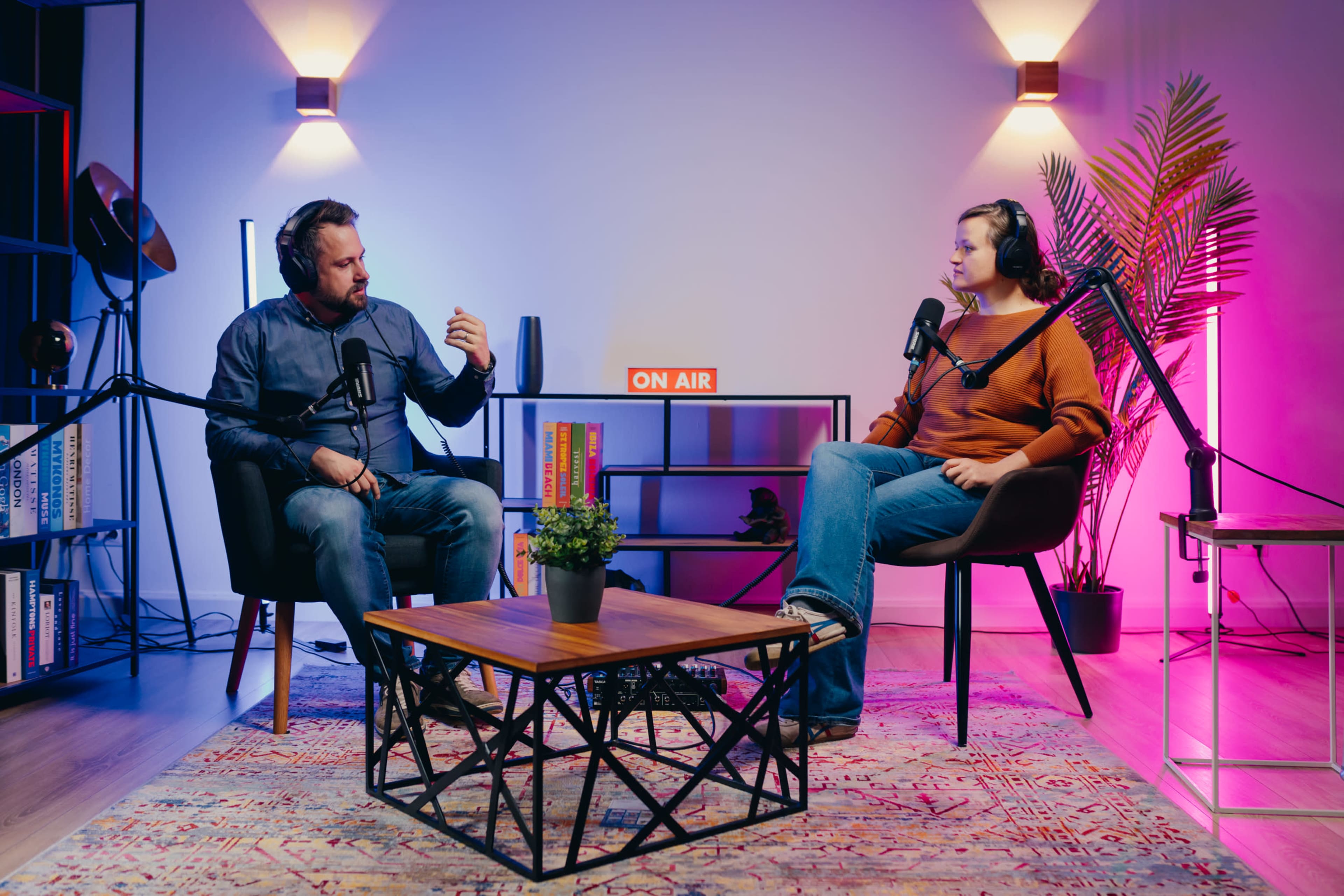 A man and a woman sit in a podcast studio, equipped with microphones and headphones, discussing in front of a "ON AIR" sign.