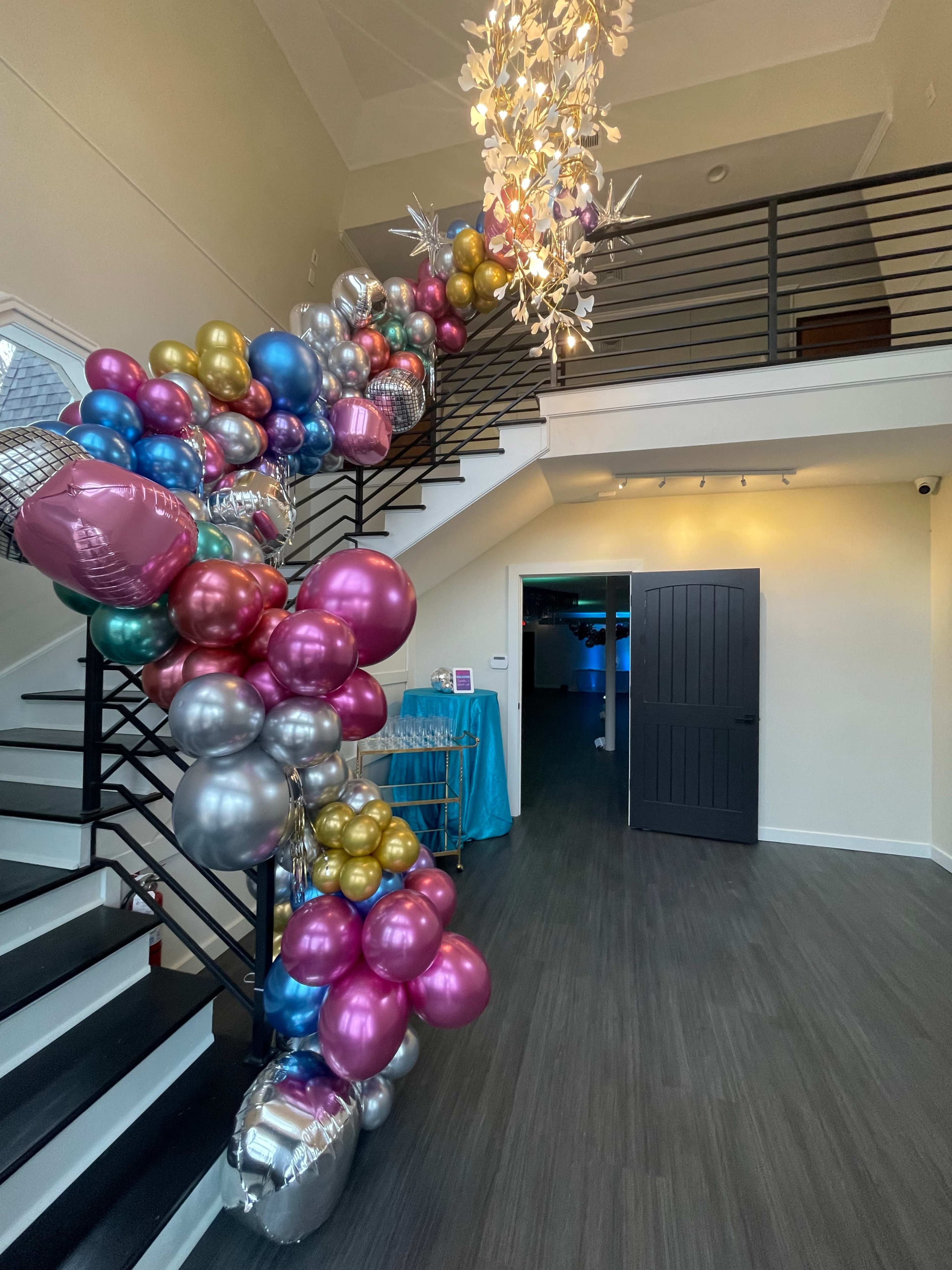 A colorful arrangement of balloons decorates the staircase in a well-lit venue, with a modern chandelier hanging above.