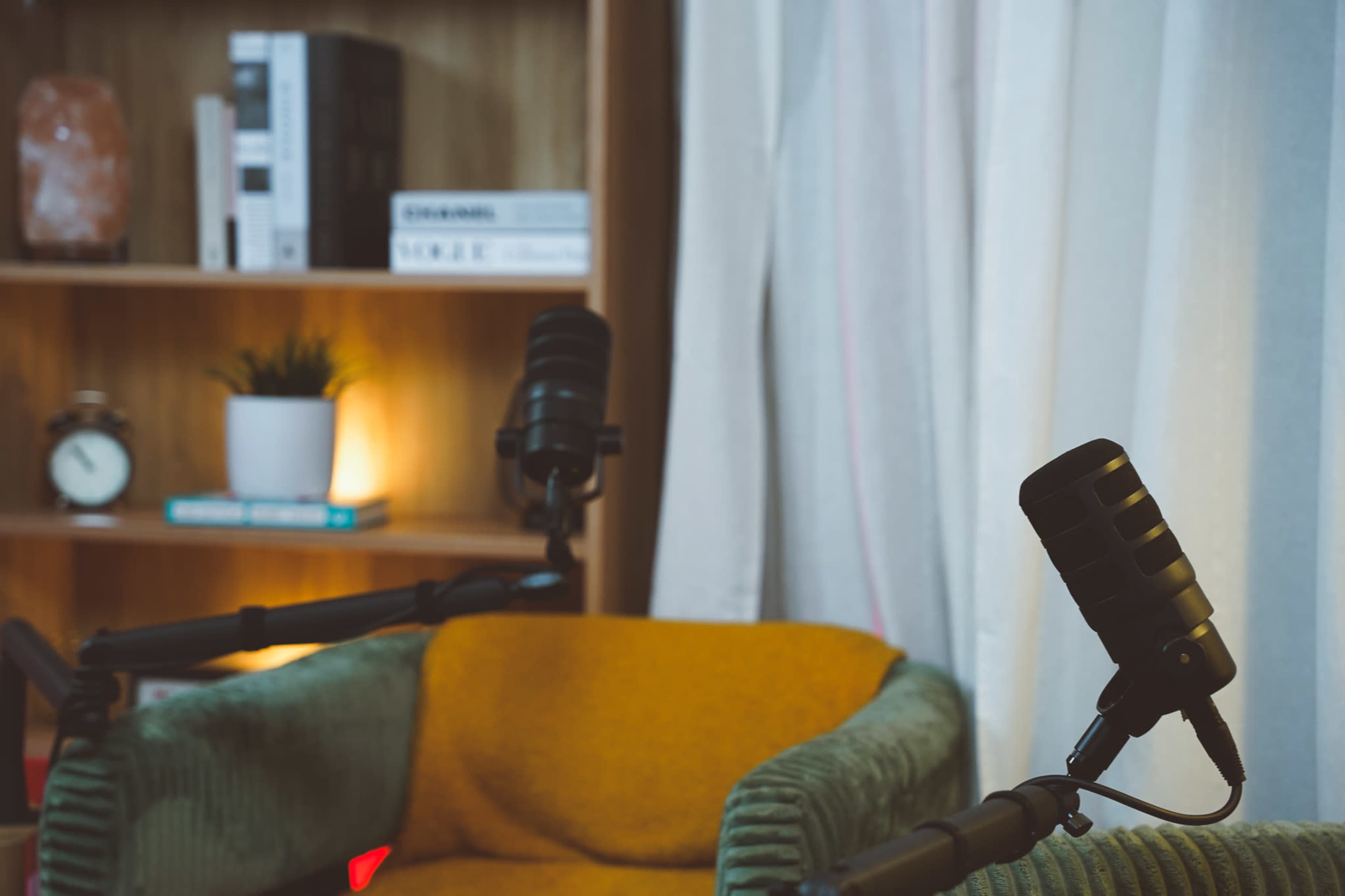 The image features a cozy seating area with two microphones positioned on stands and a bookshelf in the background, illuminated by soft lighting.
