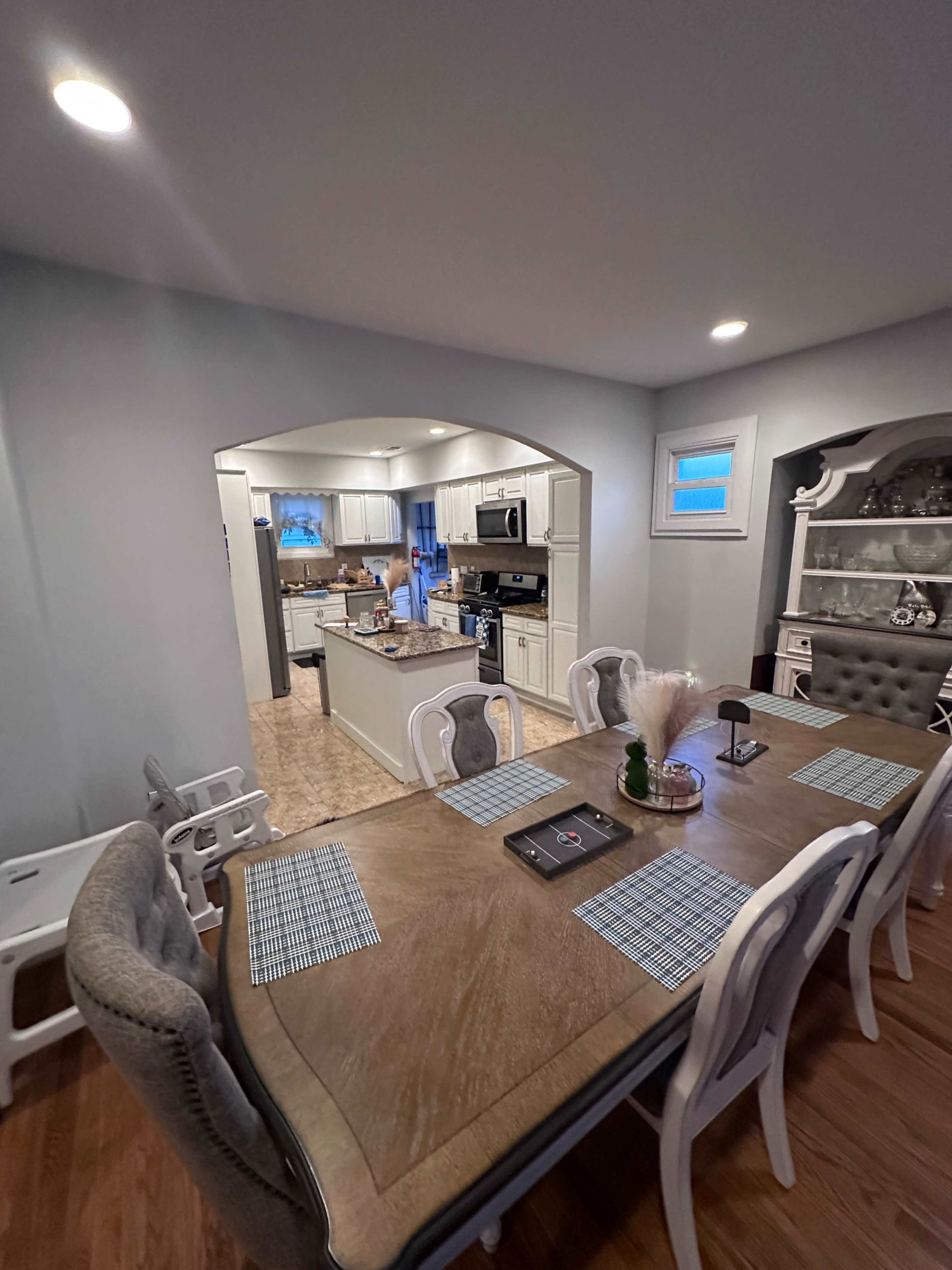 The image shows a dining area with a wooden table set for a meal, leading into a kitchen visible in the background.
