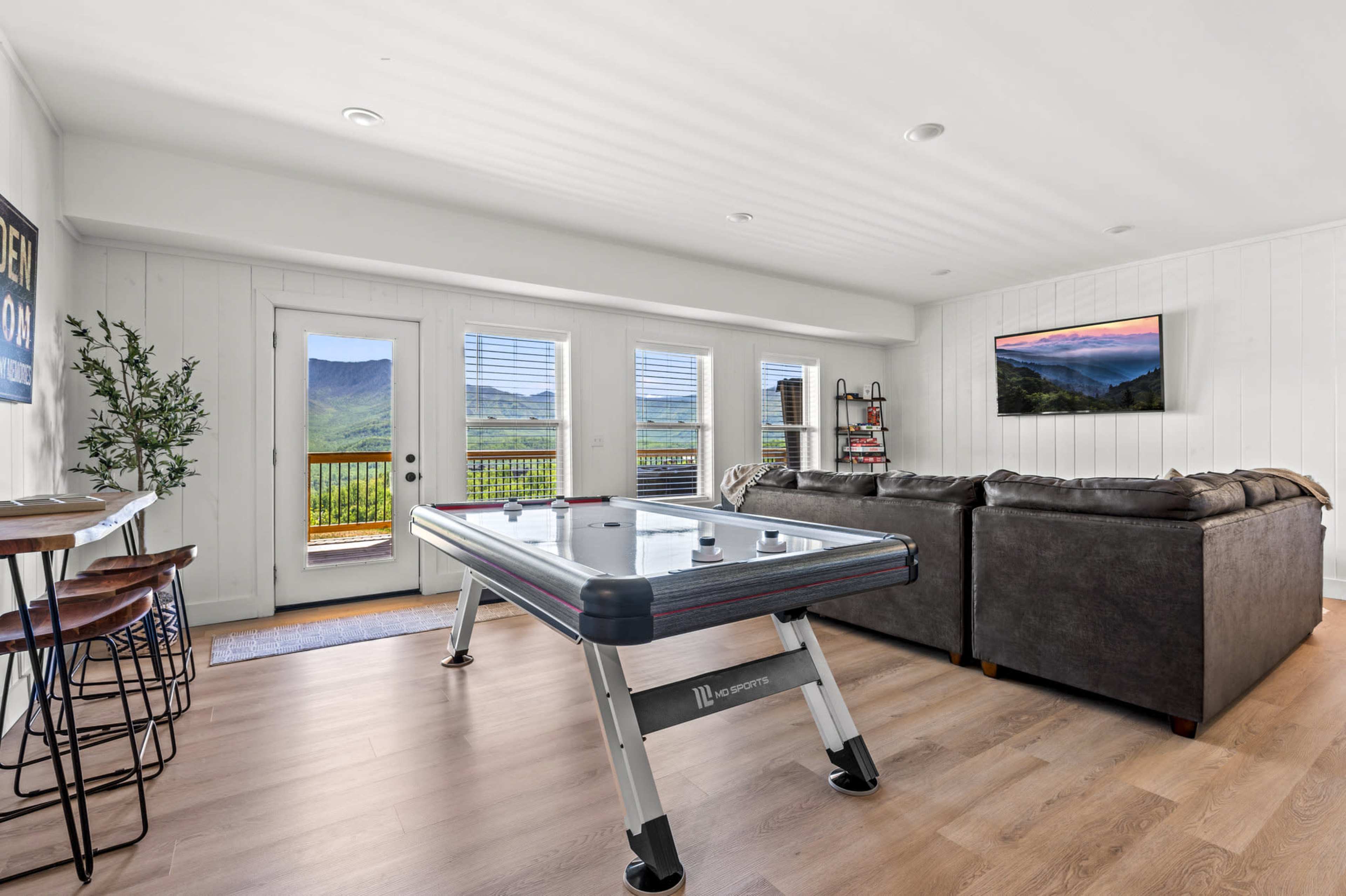 The image shows a cozy living room with a pool table, a large couch, and glass doors opening to a scenic view of mountains.