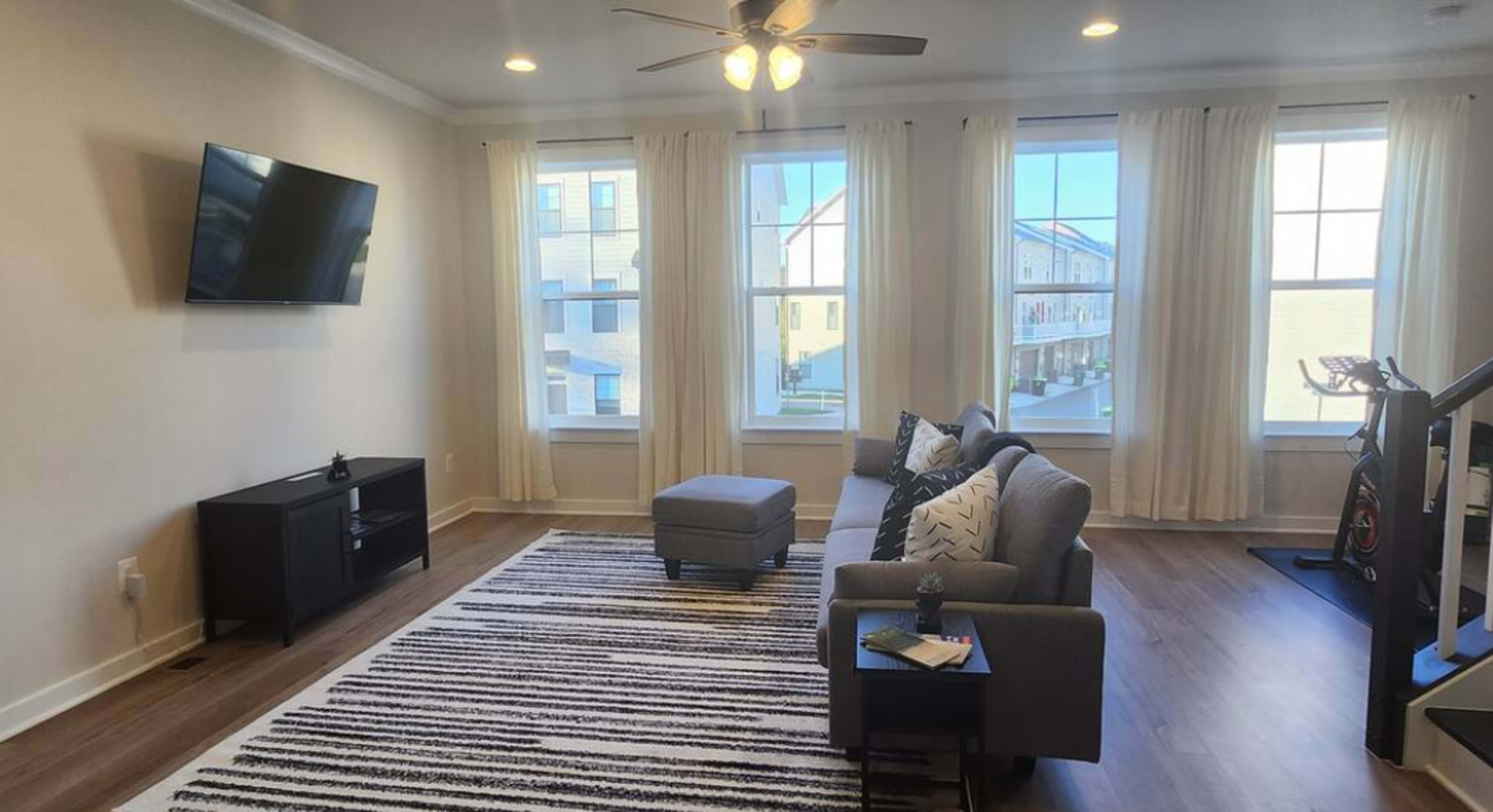 The living room features a television mounted on the wall, a gray sofa with striped accent pillows, a small ottoman, and large windows with sheer curtains.