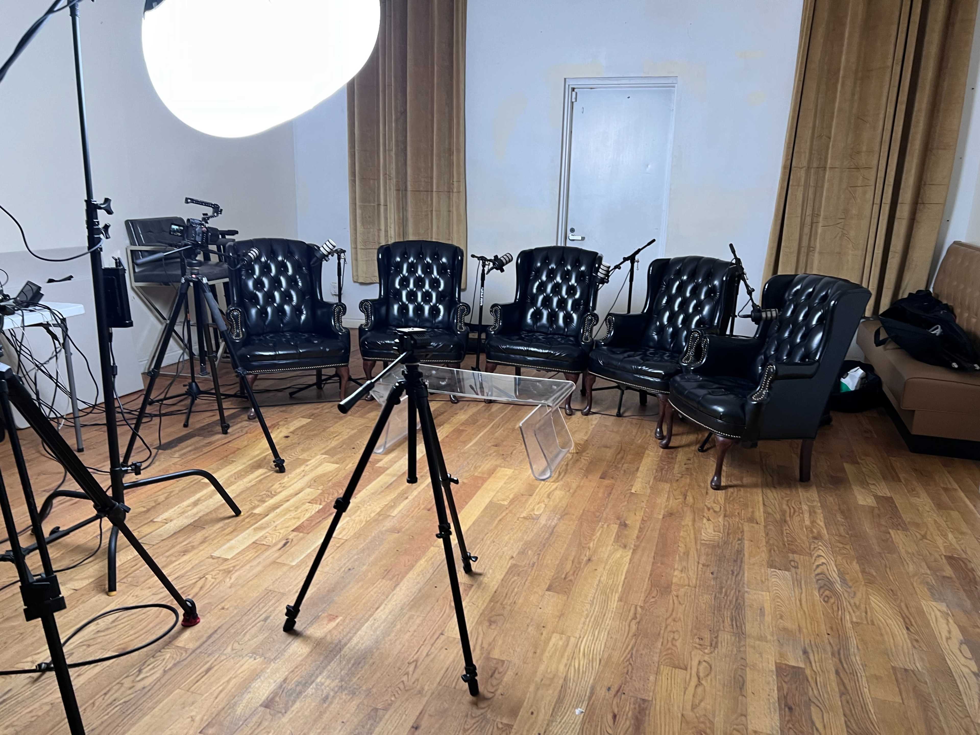 A small studio setup featuring six black leather chairs arranged in a semicircle, with cameras and lighting equipment positioned nearby on a wooden floor.