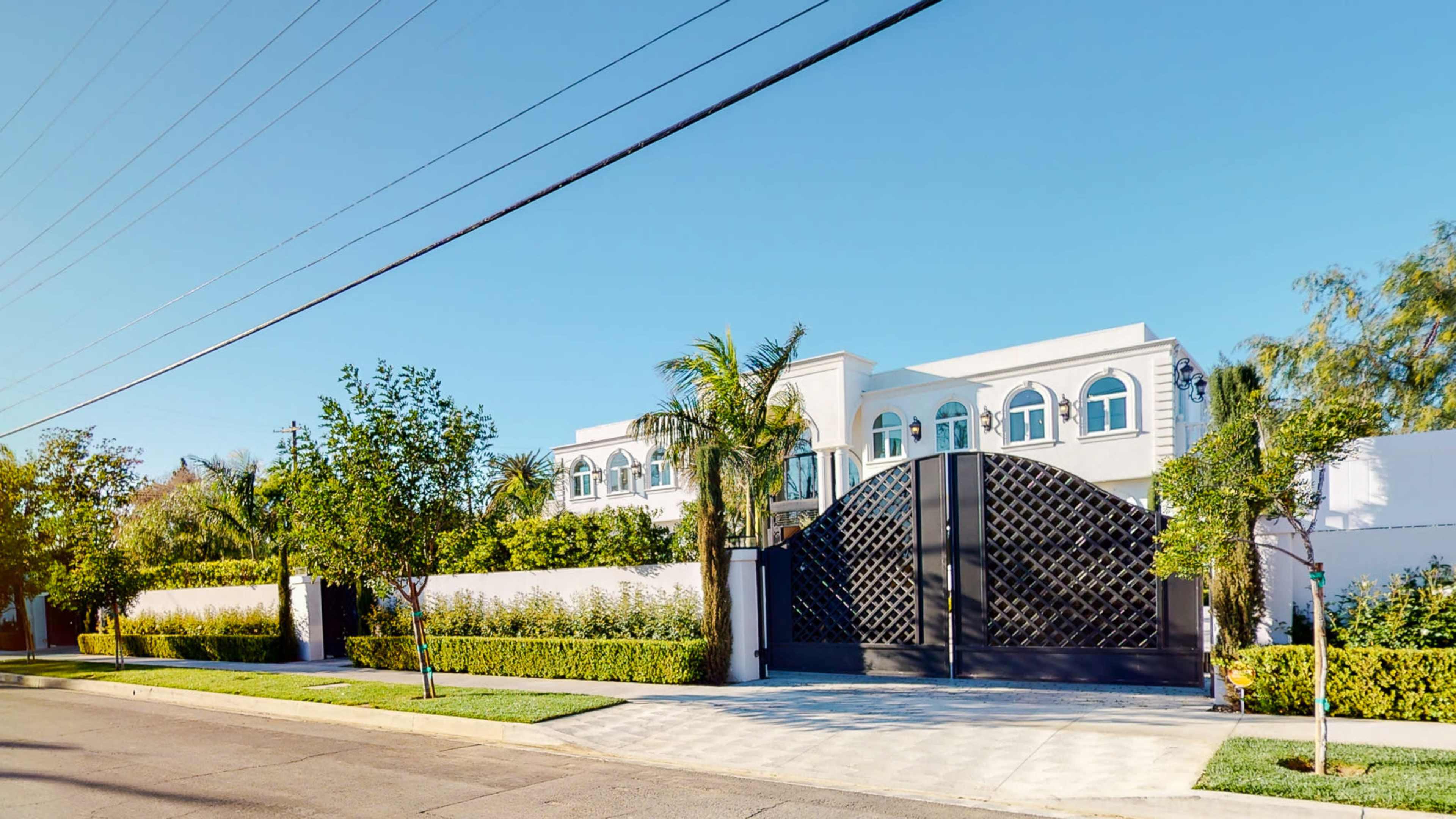 A large white house with a black gate and palm trees is surrounded by a well-maintained hedge on a sunny street.