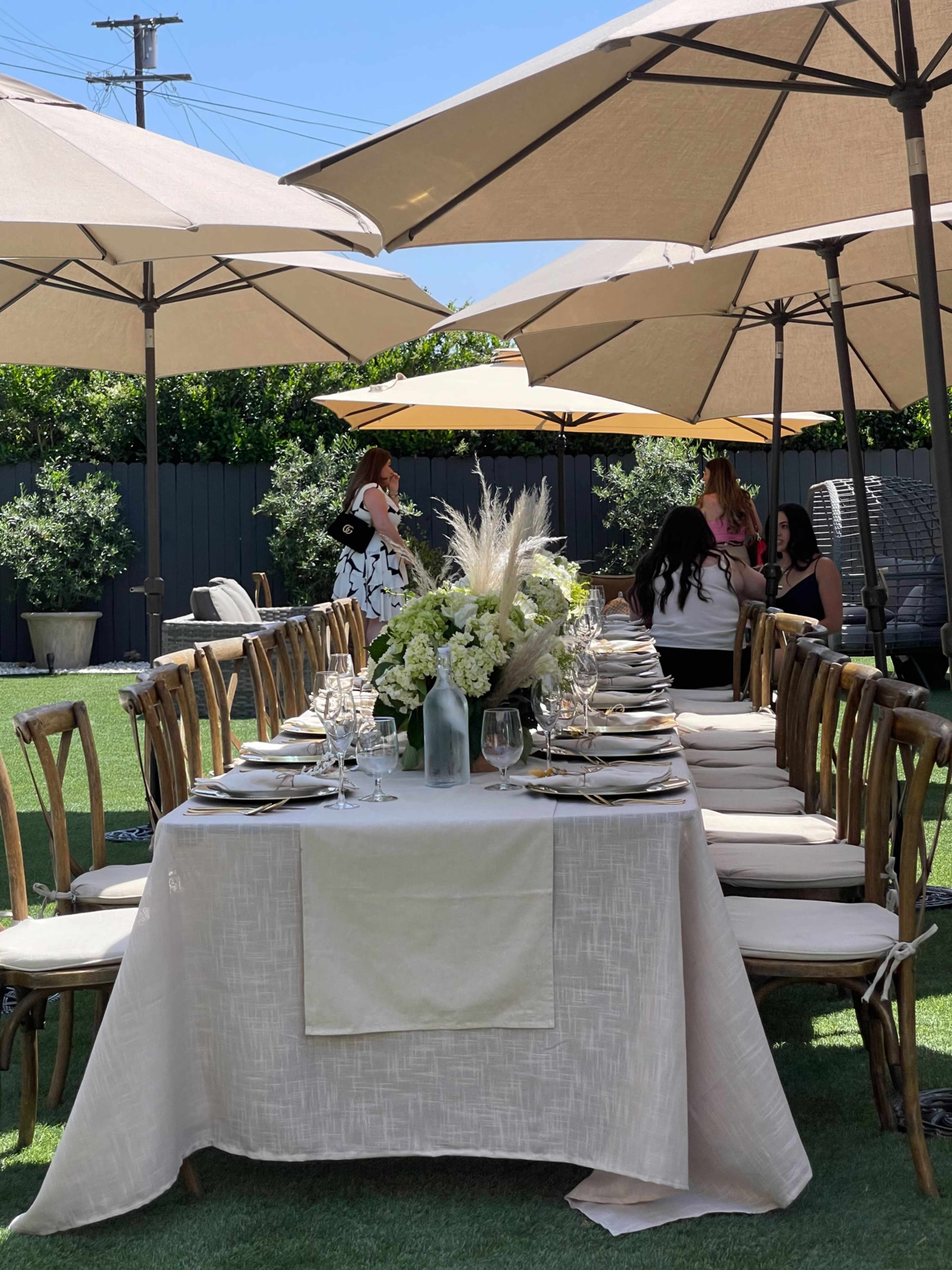 A long dining table is set for an outdoor gathering, shaded by large umbrellas, with decorative centerpieces and empty place settings.