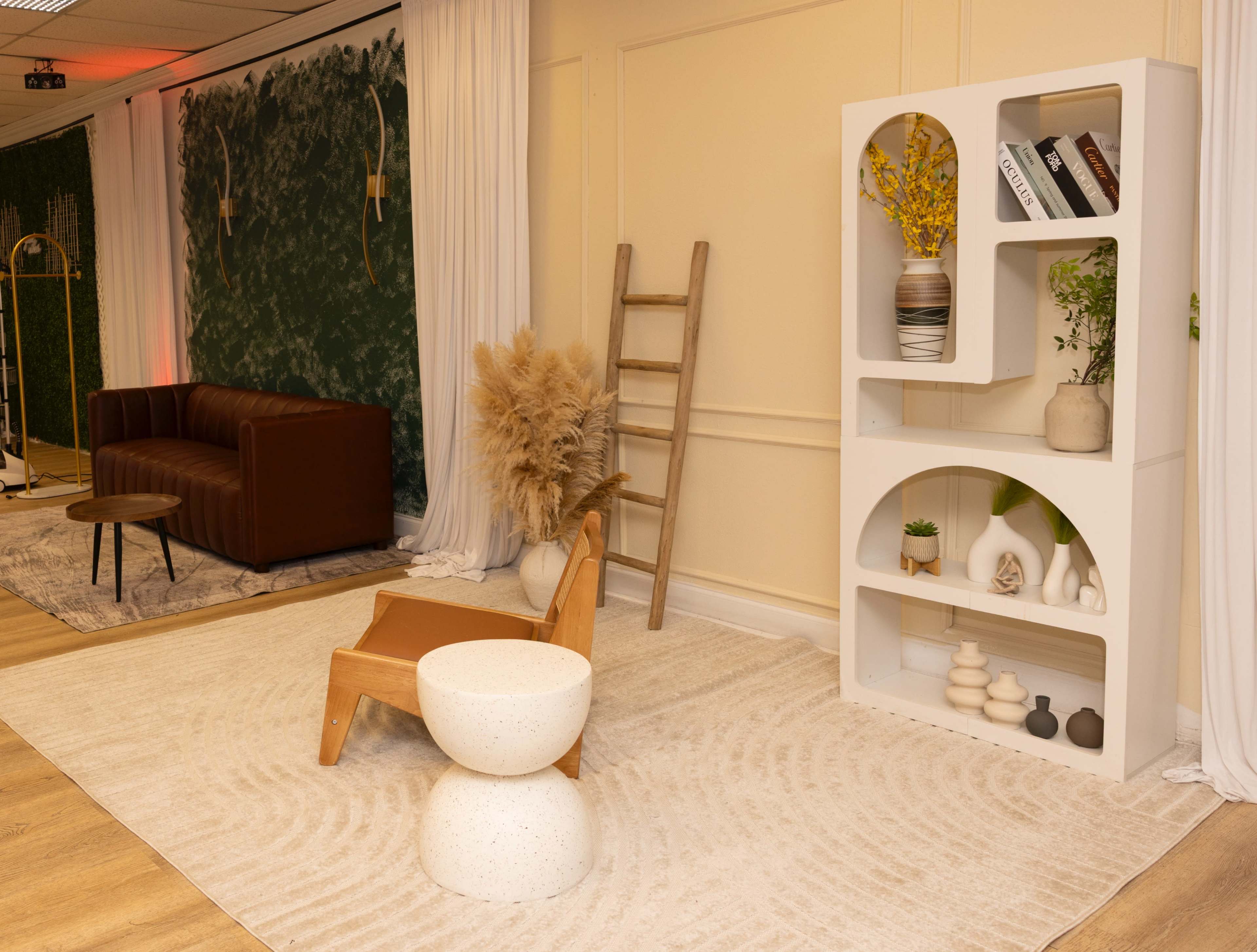 The image shows a modern interior space featuring a brown sofa, a white bookshelf with decorative items, a wooden ladder, and a round white table, all arranged on a circular patterned rug.