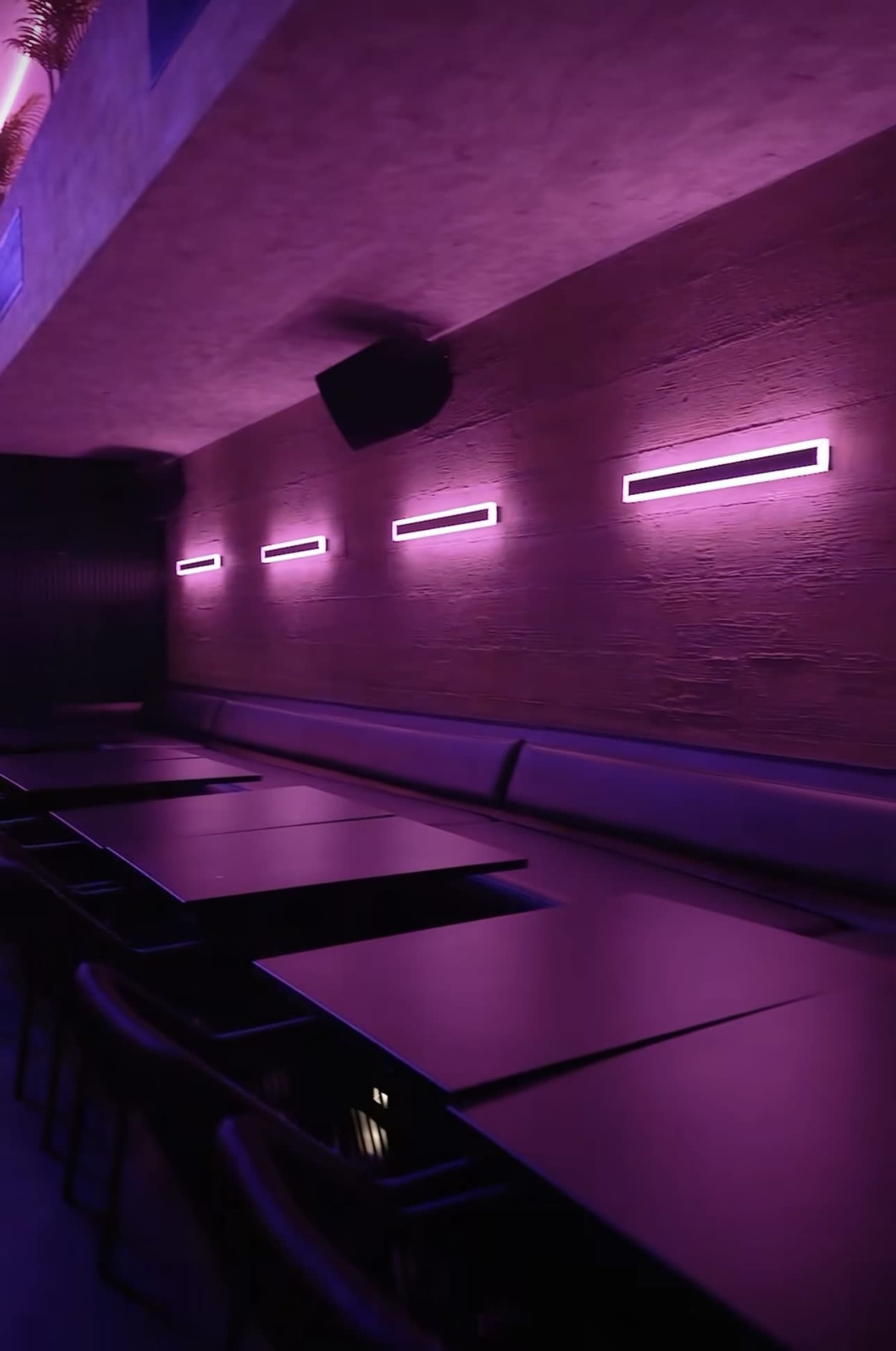 The image shows a dimly lit room with purple wall lights and a row of tables along a cushioned bench.