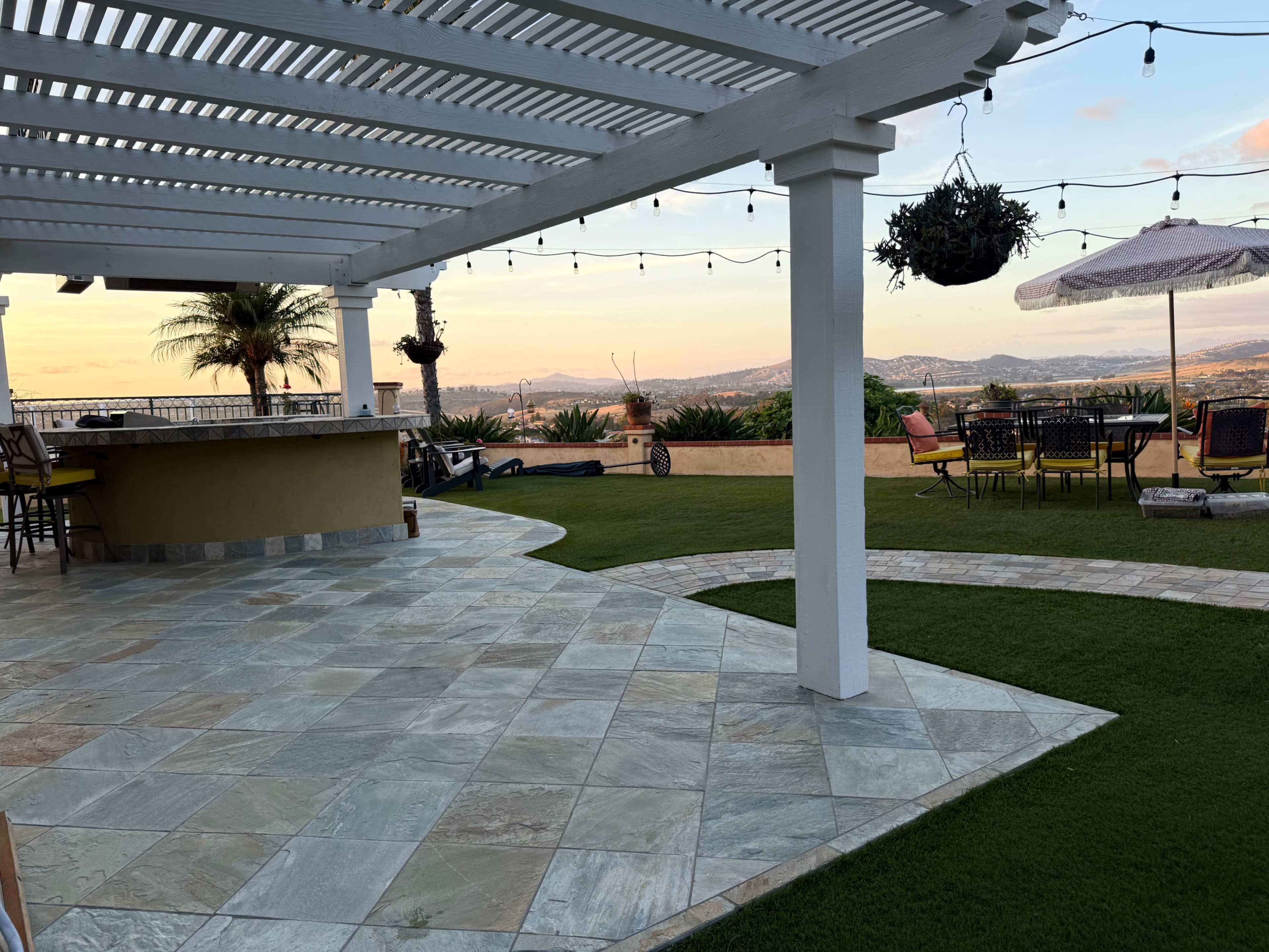 The image features a patio area with stone flooring, shaded by a lattice pergola, overlooking a landscape at sunset adorned with palm trees and outdoor seating.