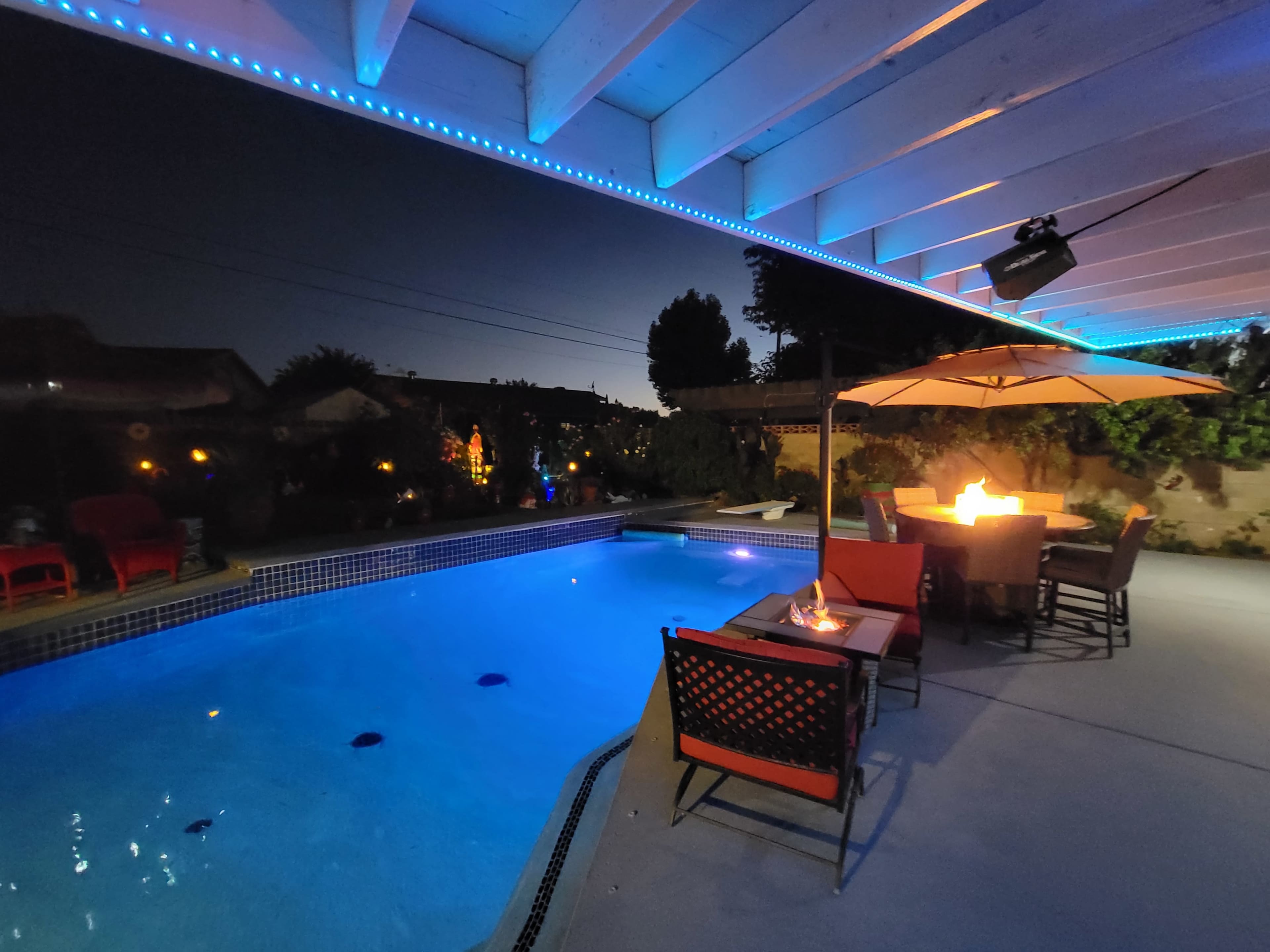A backyard pool area illuminated by blue lights, with a patio dining set and a fire pit under a covered structure at dusk.