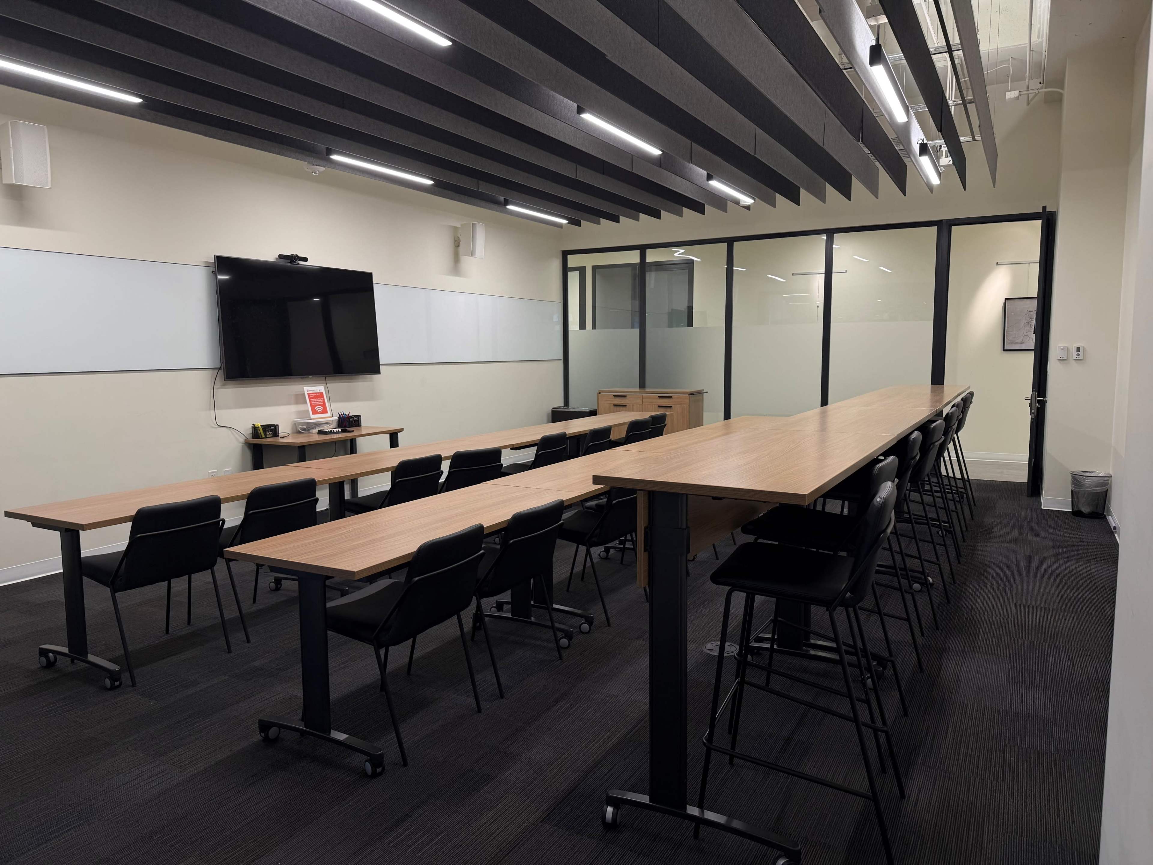 The image shows a modern conference room featuring a long table with high-backed chairs, a large screen for presentations, and glass walls that separate it from an adjacent space.