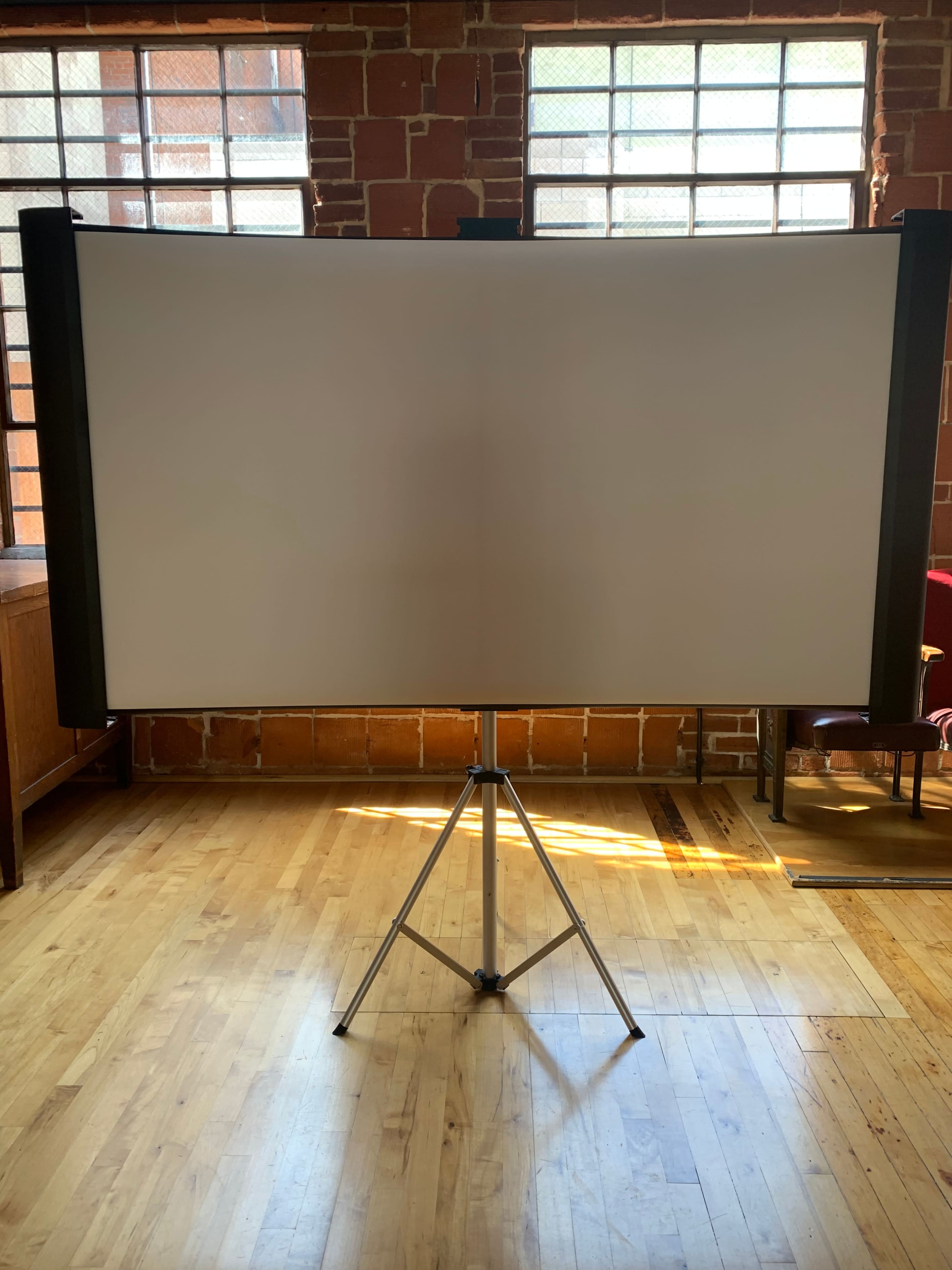 A large, blank projection screen set up on a tripod in a room with wooden floors and brick walls.