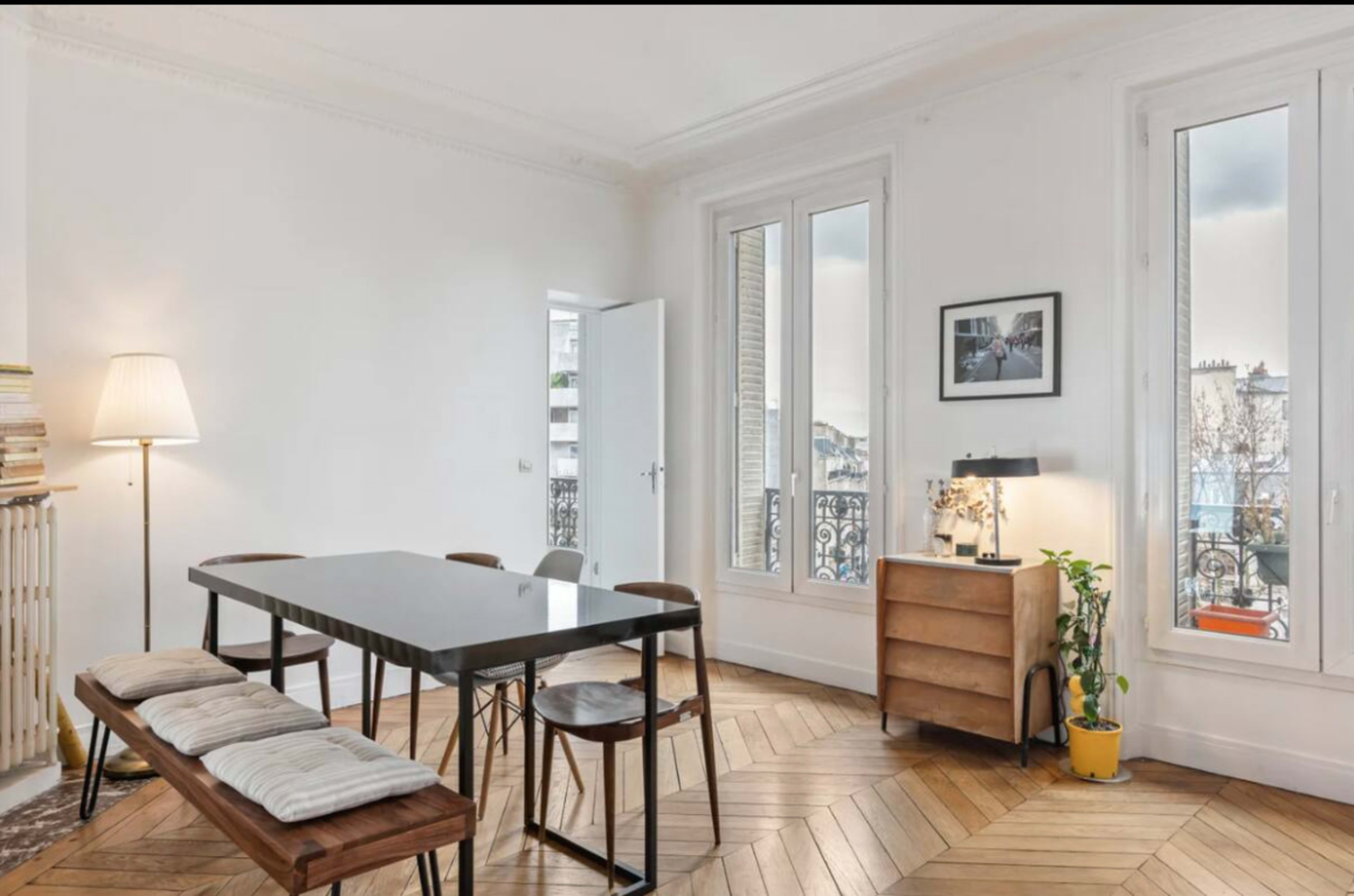 The image depicts a bright, modern dining area featuring a black table, wooden benches, and a light wood dresser next to large windows with a balcony view.