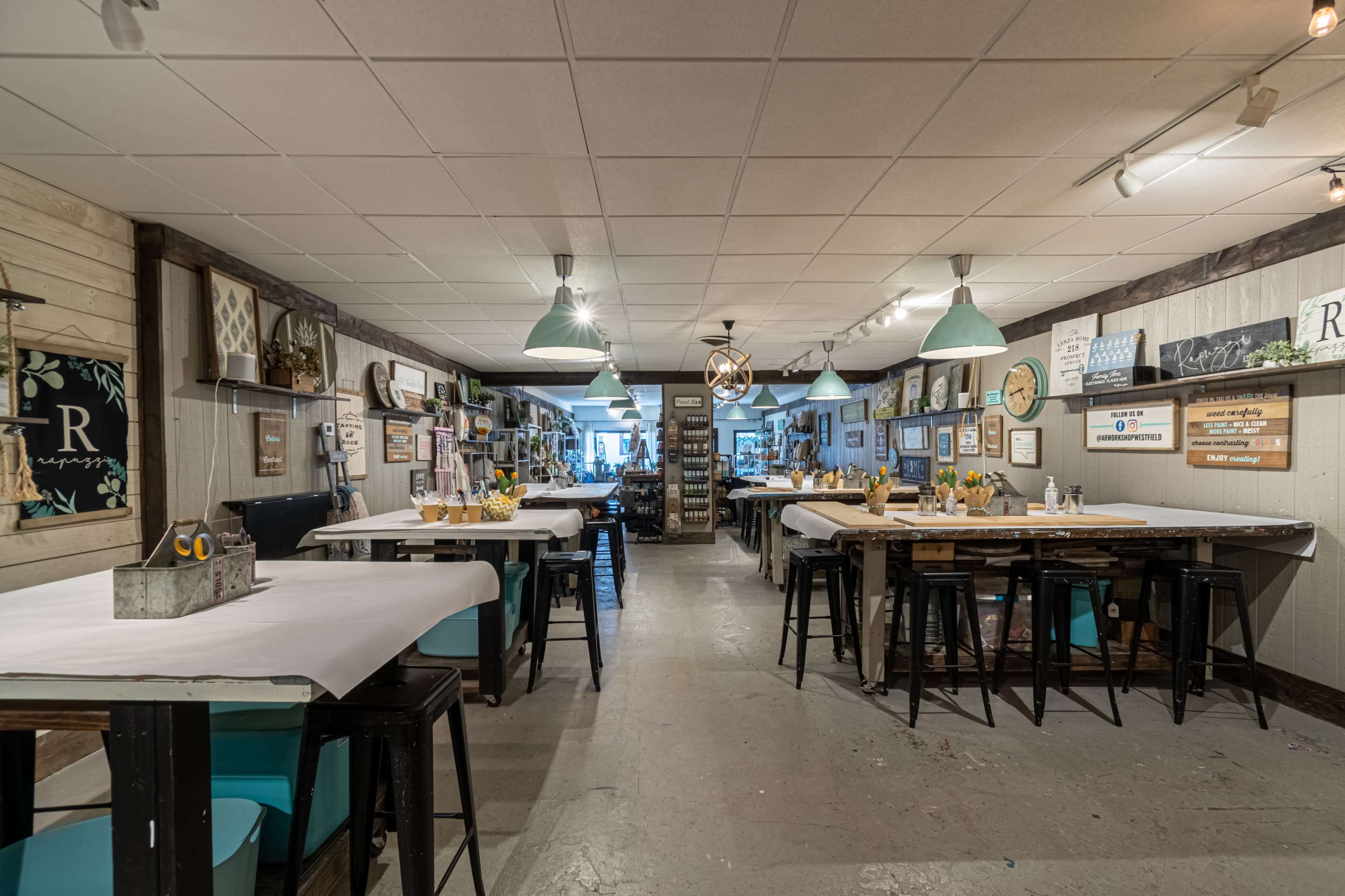 A spacious craft studio with several tables arranged along the walls, each adorned with flowers and art supplies, and various artworks hung on the walls.