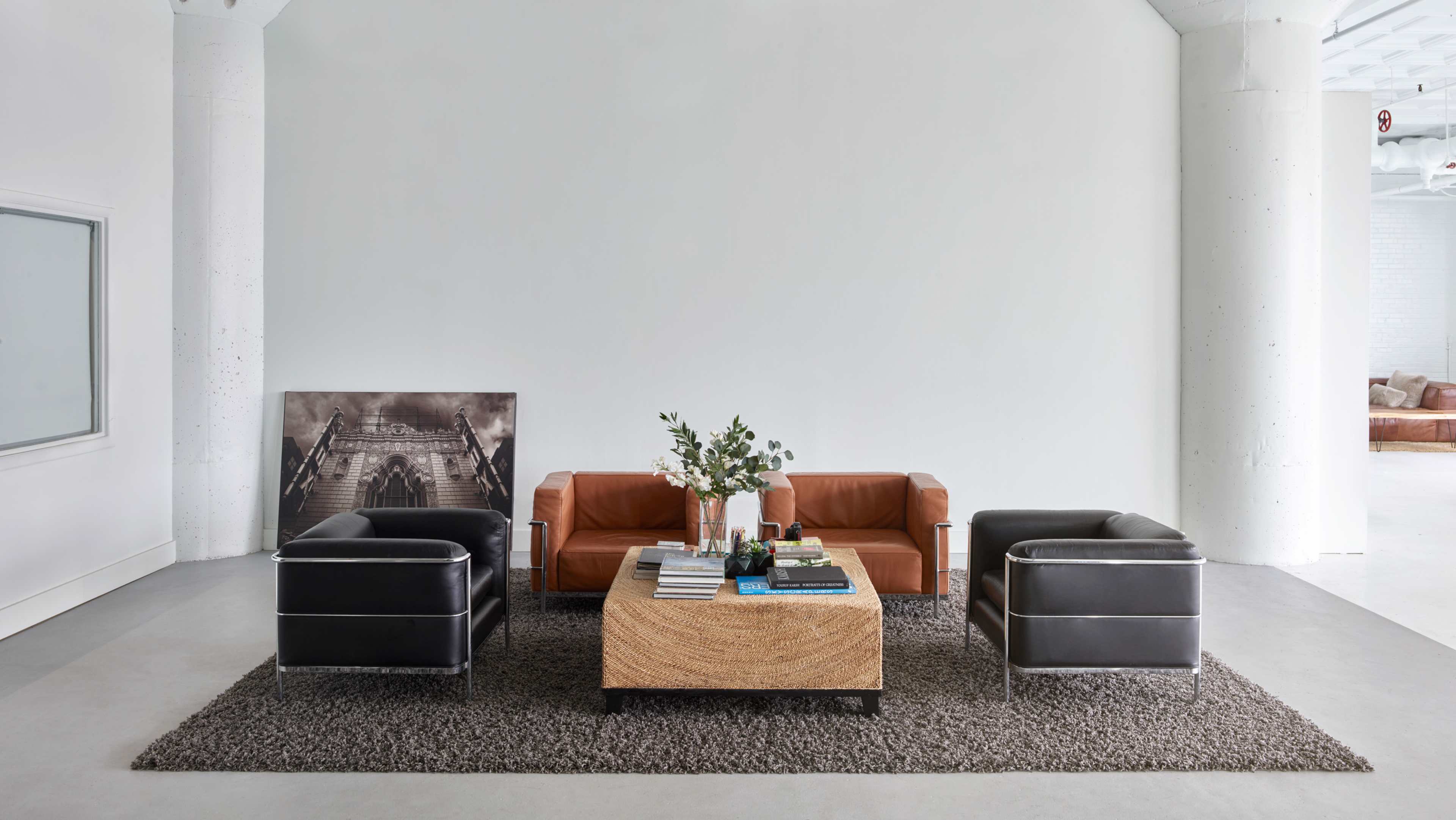 The image features a modern living room with two brown leather sofas, two black armchairs, a wooden coffee table, and a large wall art piece, all set against a light-colored wall.