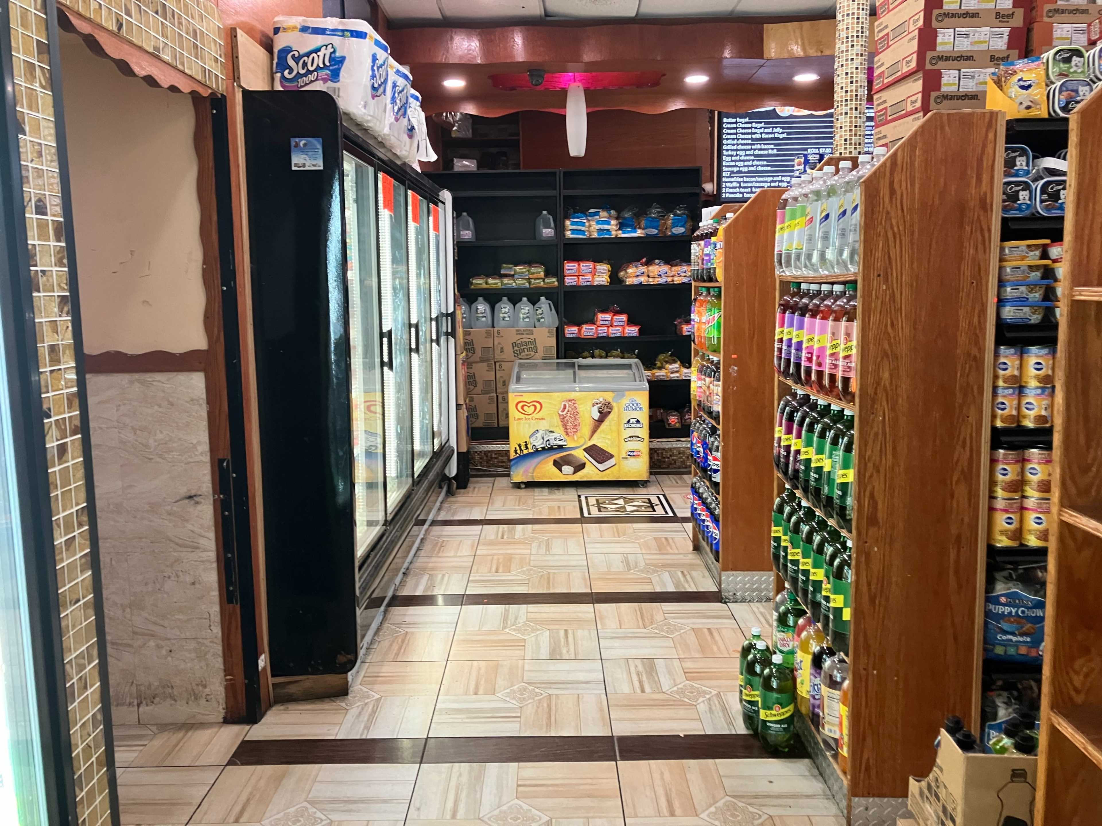 An aisle in a convenience store with shelves of beverages and snacks on either side, leading to a wall of refrigerated cases.