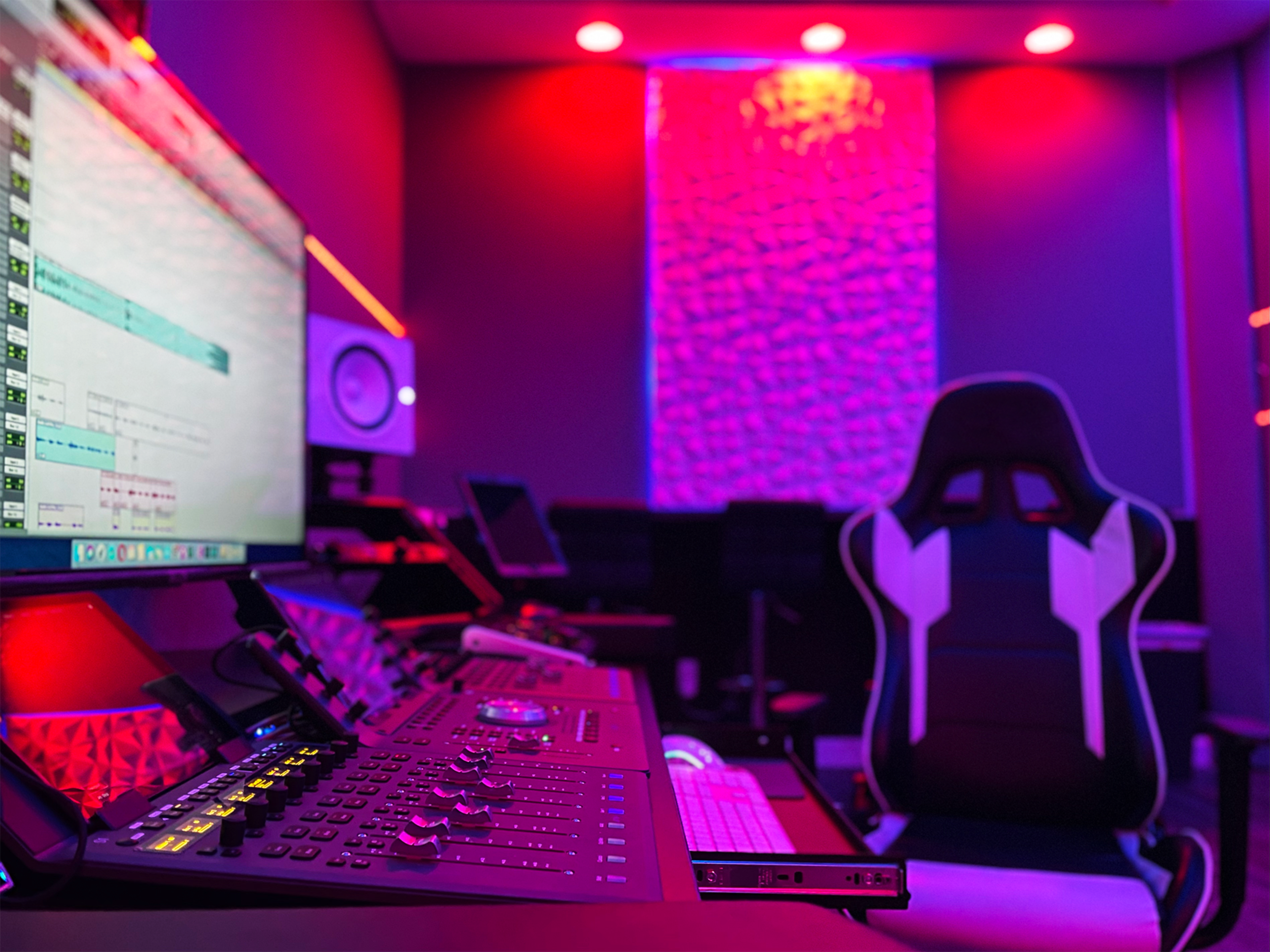 A music studio featuring a computer monitor displaying audio software, a control panel with various knobs and buttons, and a gaming-style chair under colorful lighting.