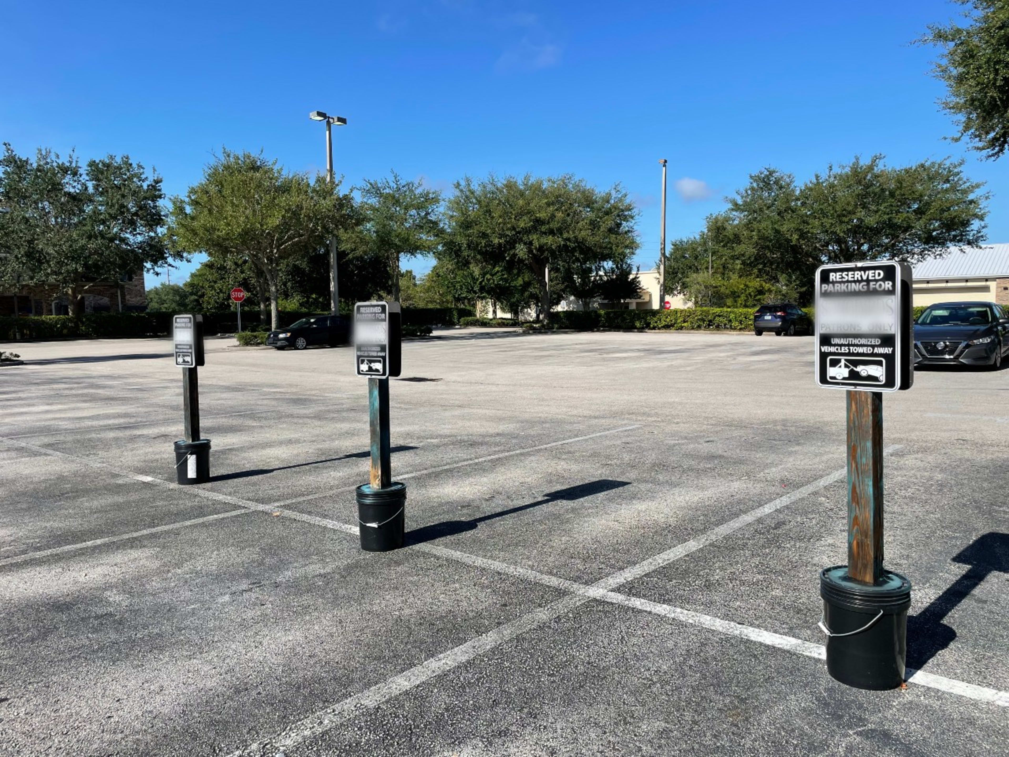 Five parking meters are positioned in a mostly empty parking lot surrounded by trees under a clear blue sky.