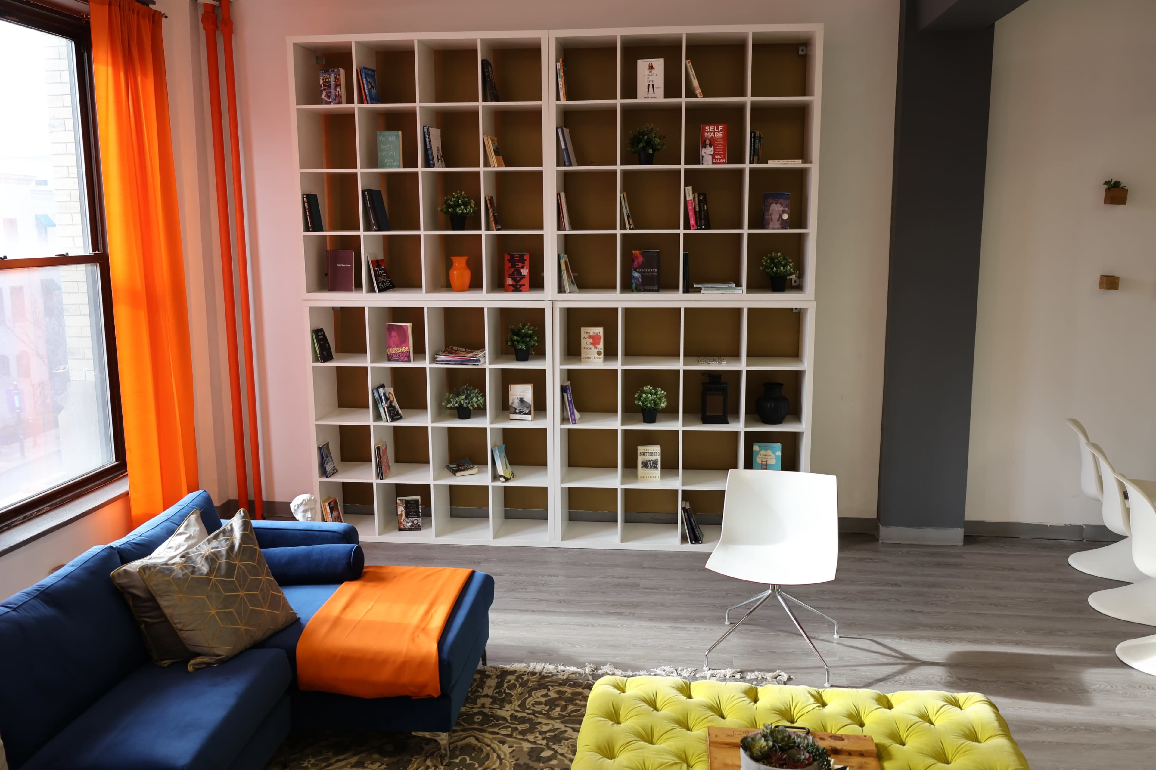 A modern living space features a blue sofa with an orange throw, a bright yellow ottoman, and a white shelving unit filled with books and decorative items.