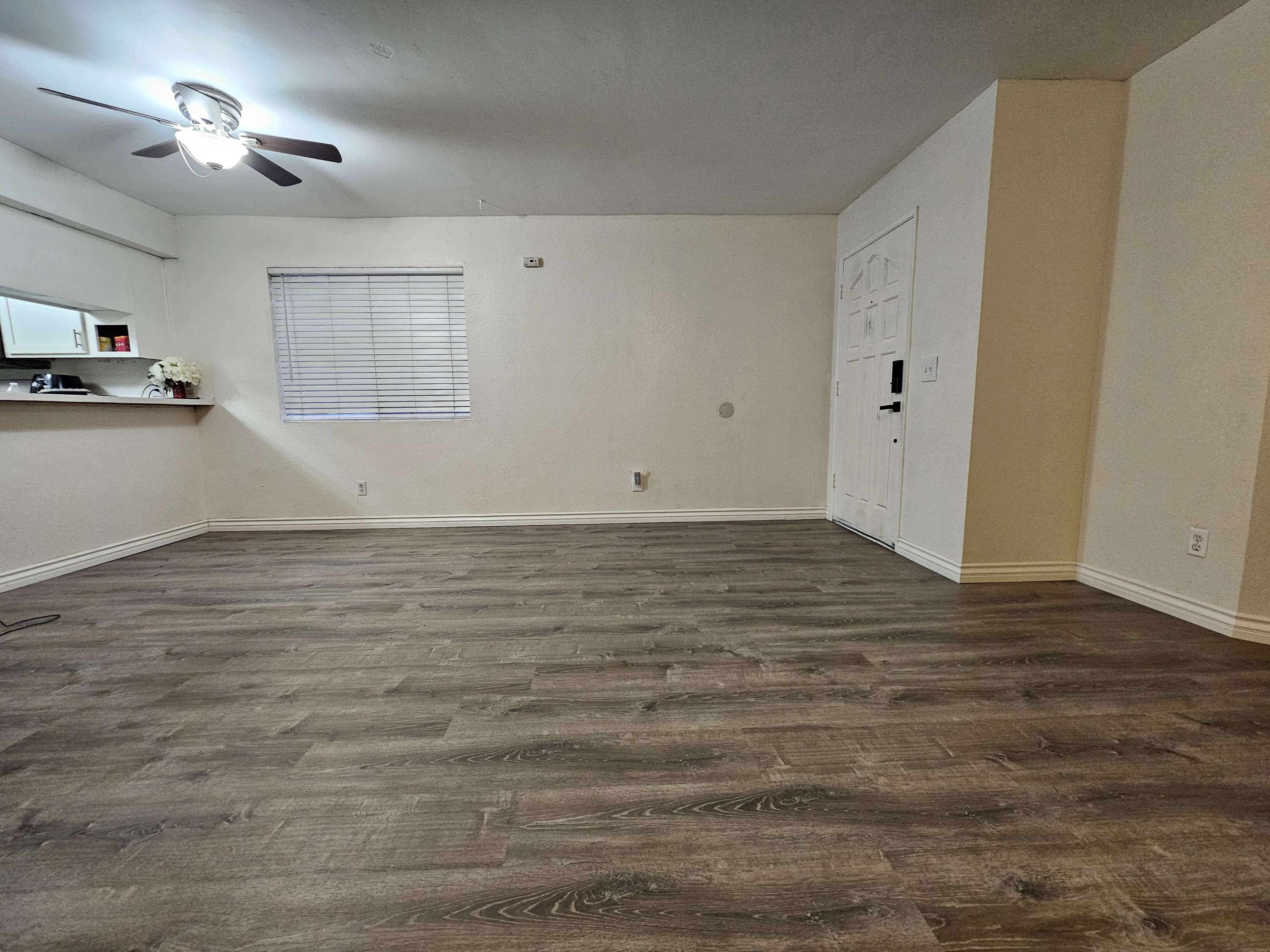 A spacious, empty living room with laminate flooring and a ceiling fan, featuring a window and an entrance door.