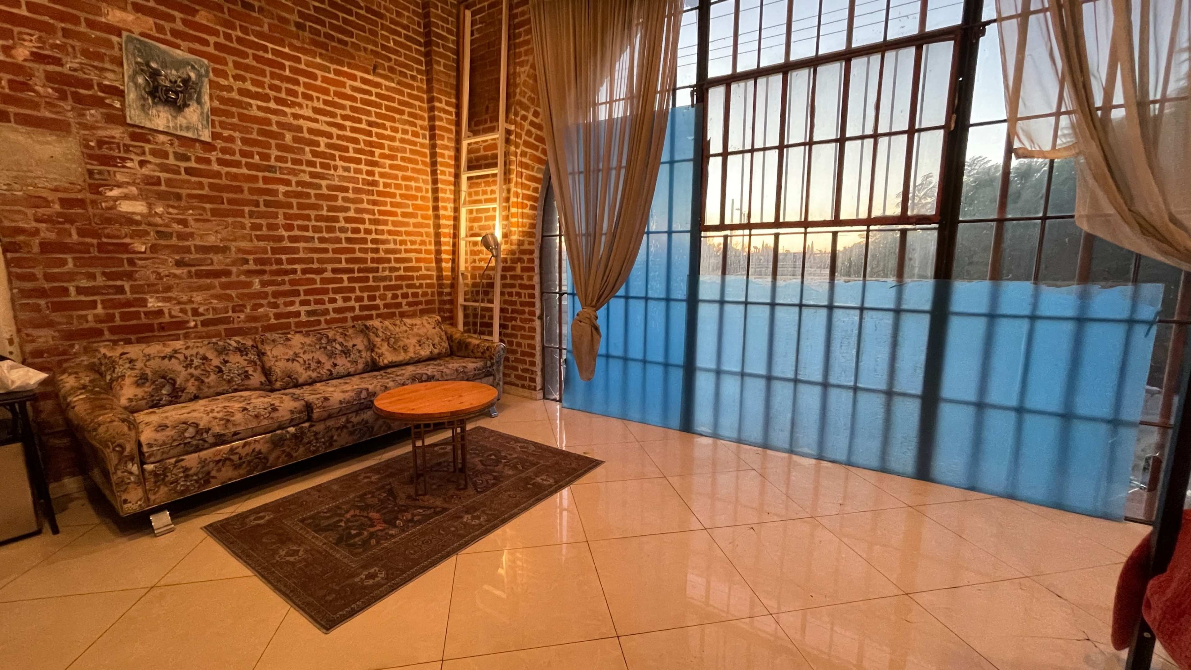 The image shows a cozy room with exposed brick walls, a floral couch, a small wooden table, and large windows draped with sheer curtains, revealing a sunset outside.
