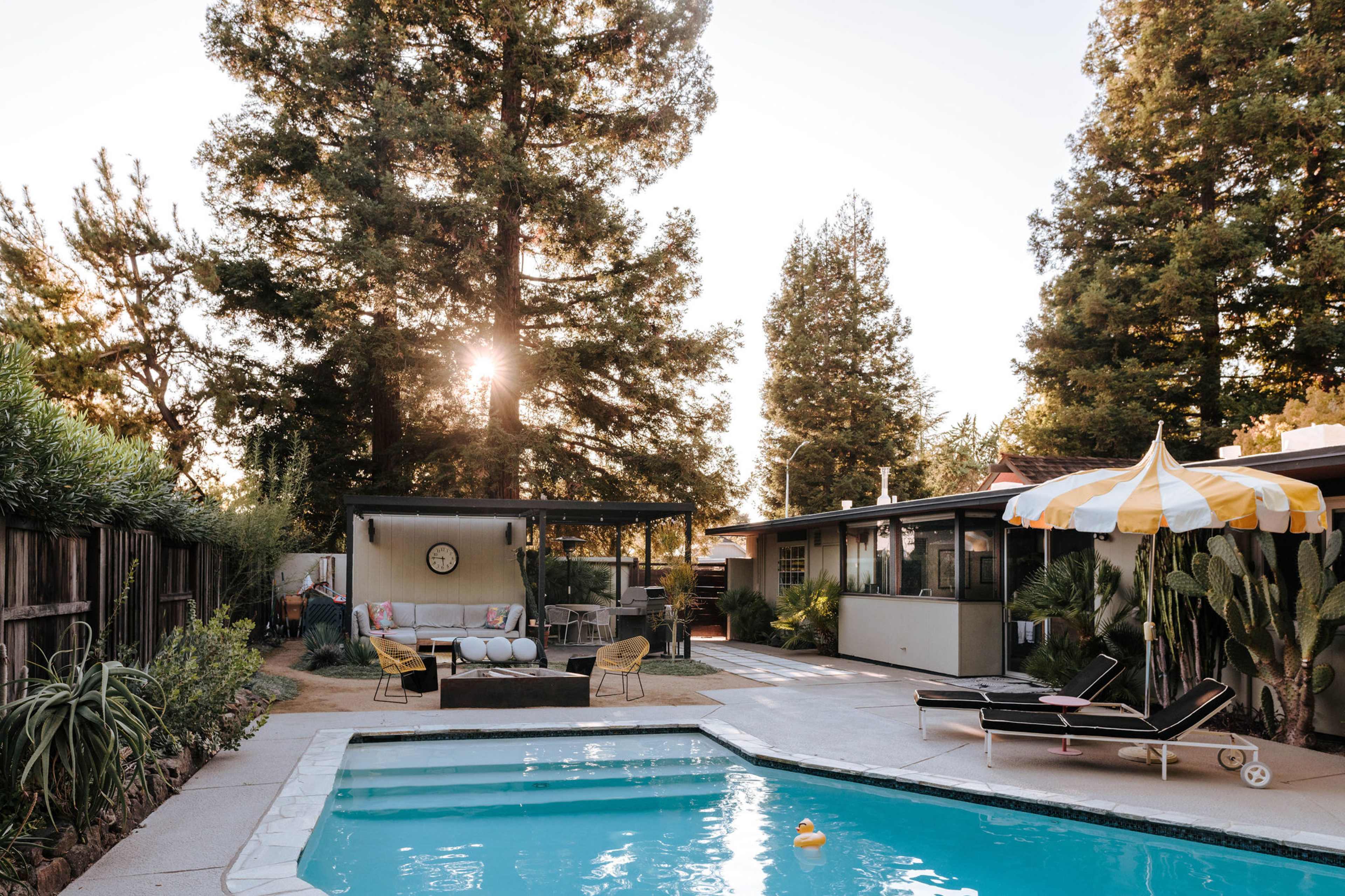 A swimming pool with a yellow-striped umbrella is surrounded by a landscaped yard and a modern outdoor seating area.