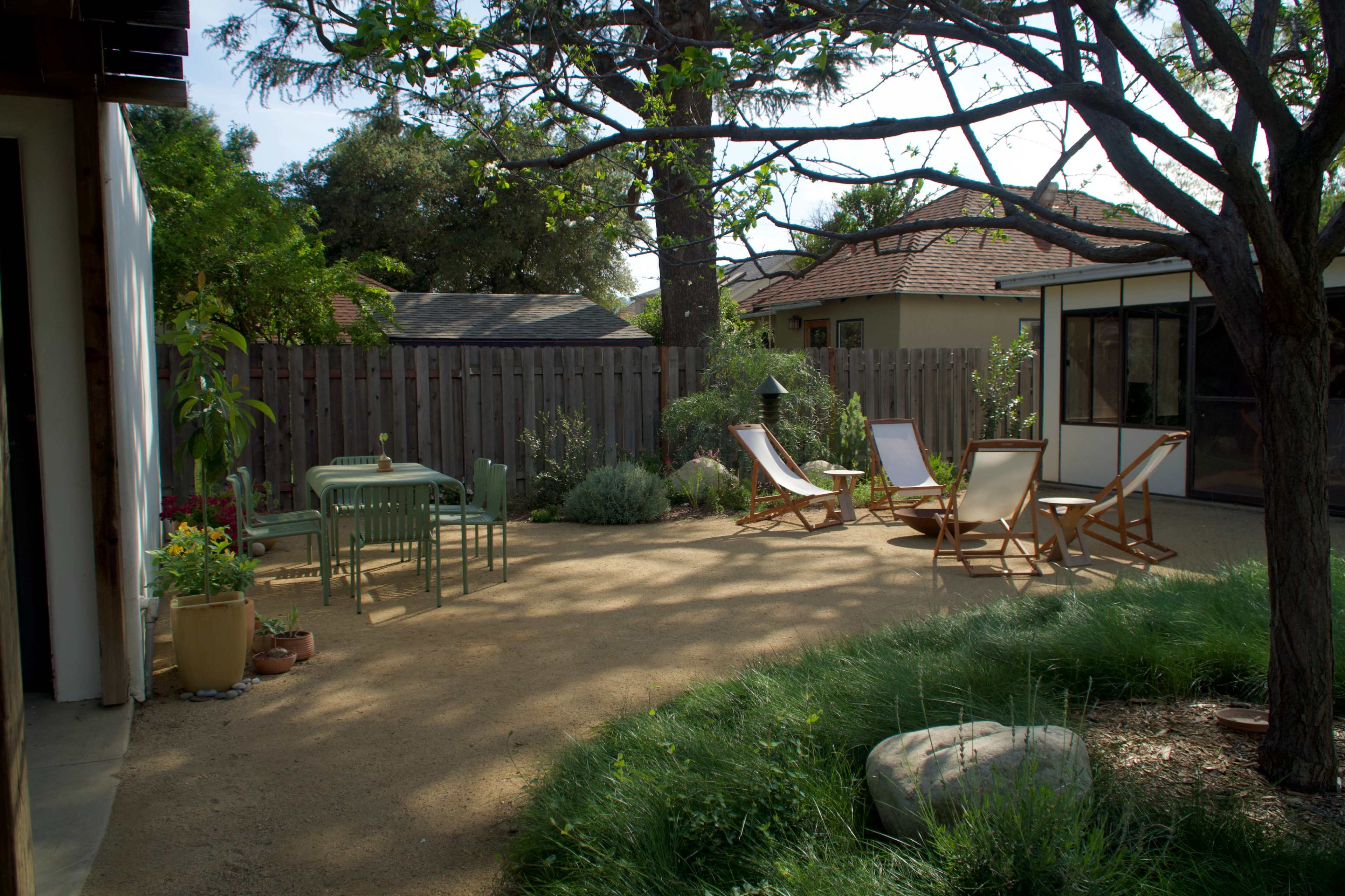 The scene shows a spacious backyard with a gravel patio featuring a table and chairs, two wooden lounge chairs, and various plants surrounded by a wooden fence and trees.