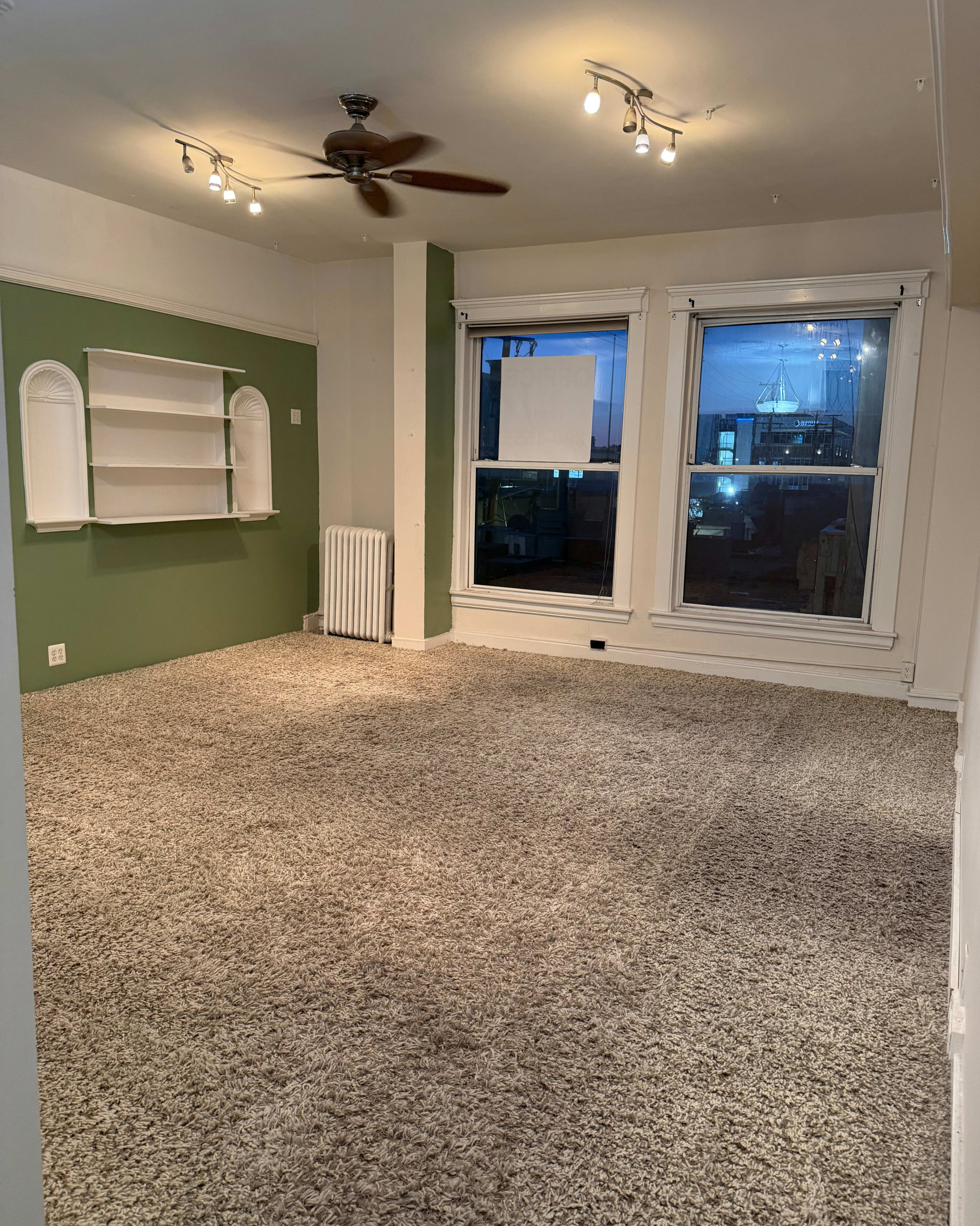 The room features light-colored walls, a textured carpet, a ceiling fan, a green accent wall with built-in shelves, and large windows with a view of the outdoors.