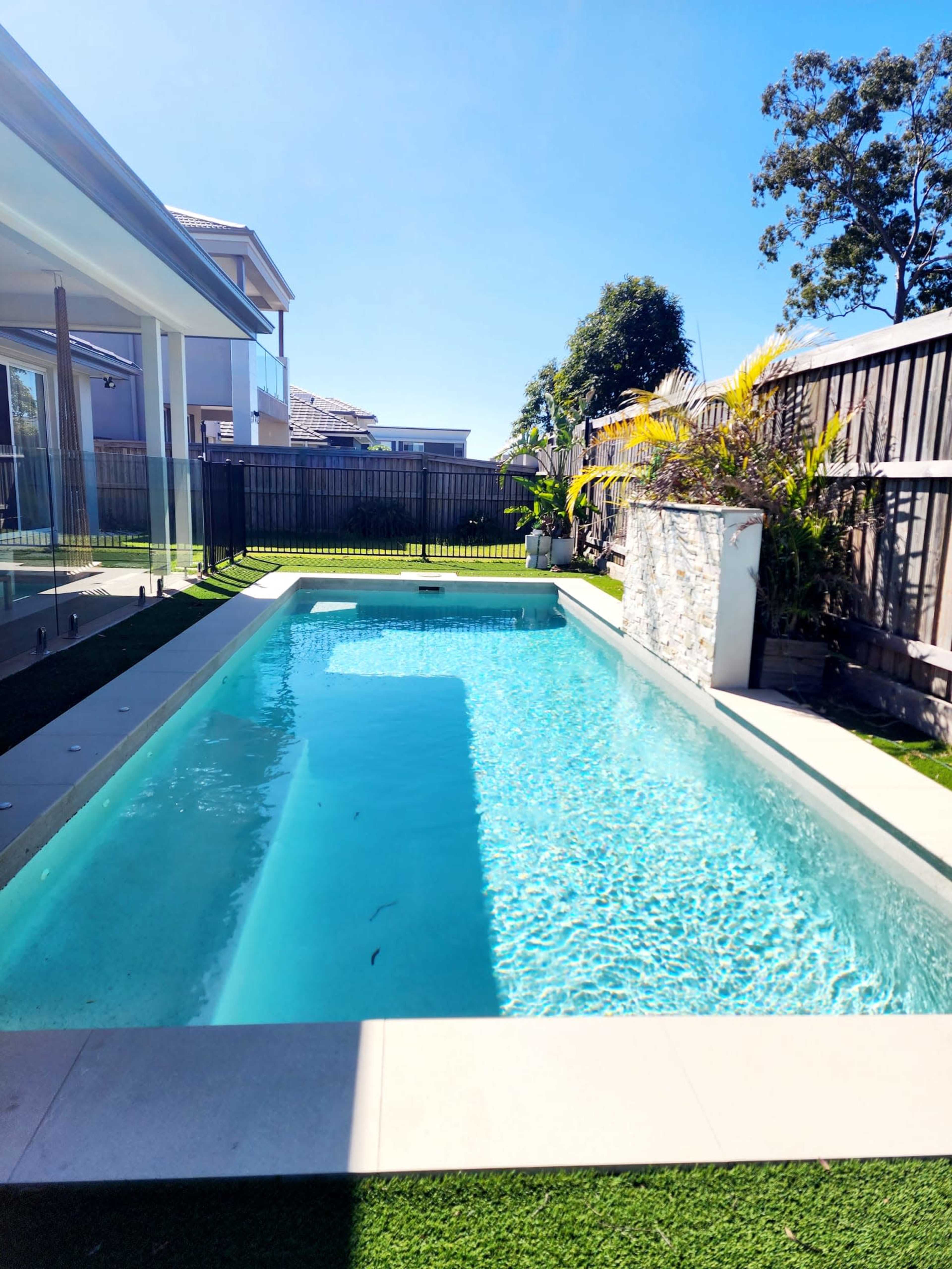 A clear swimming pool surrounded by landscaped greenery and a wooden fence.