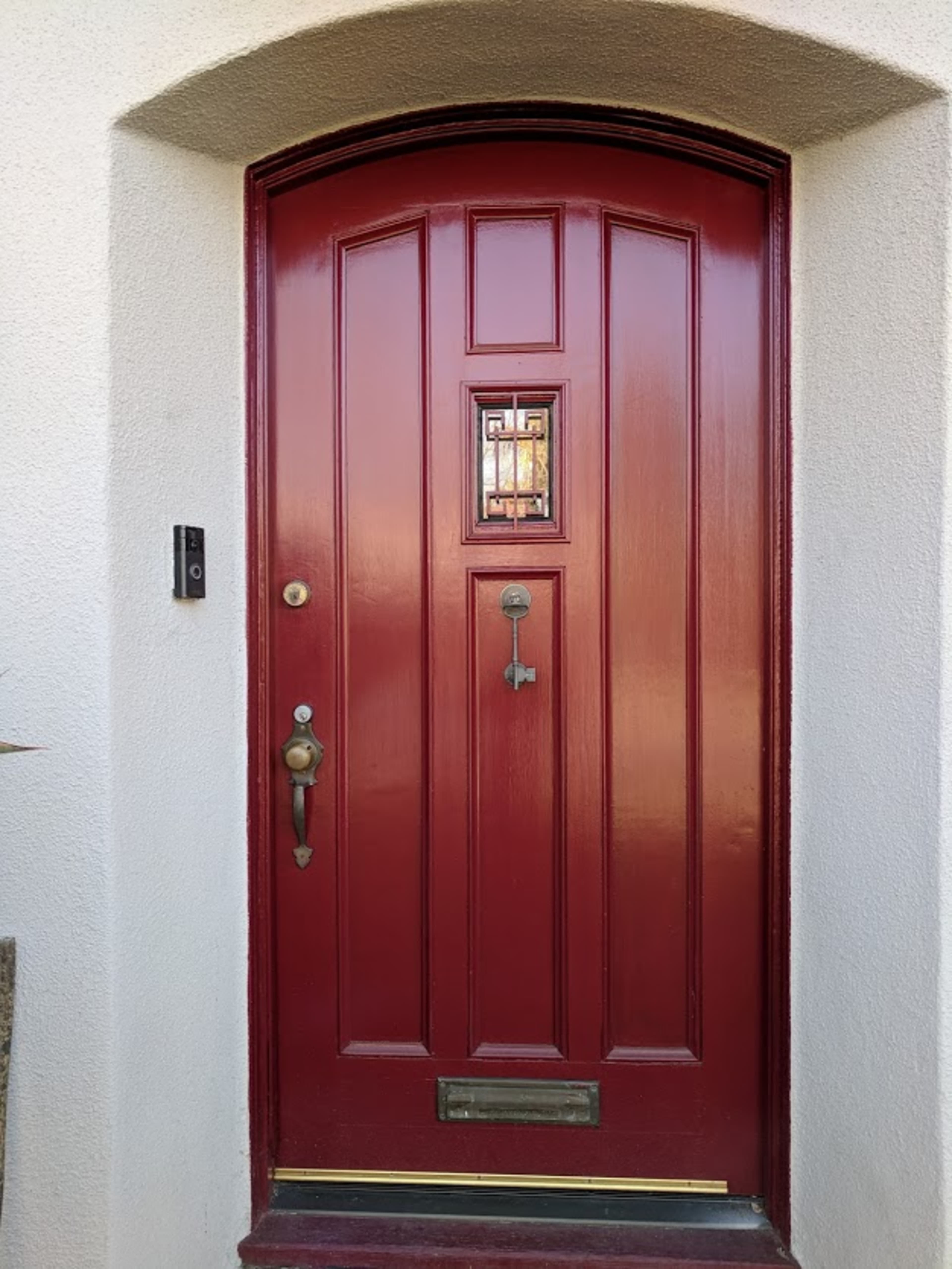 A red wooden door with a decorative window and a brass doorknob, set into a white wall.