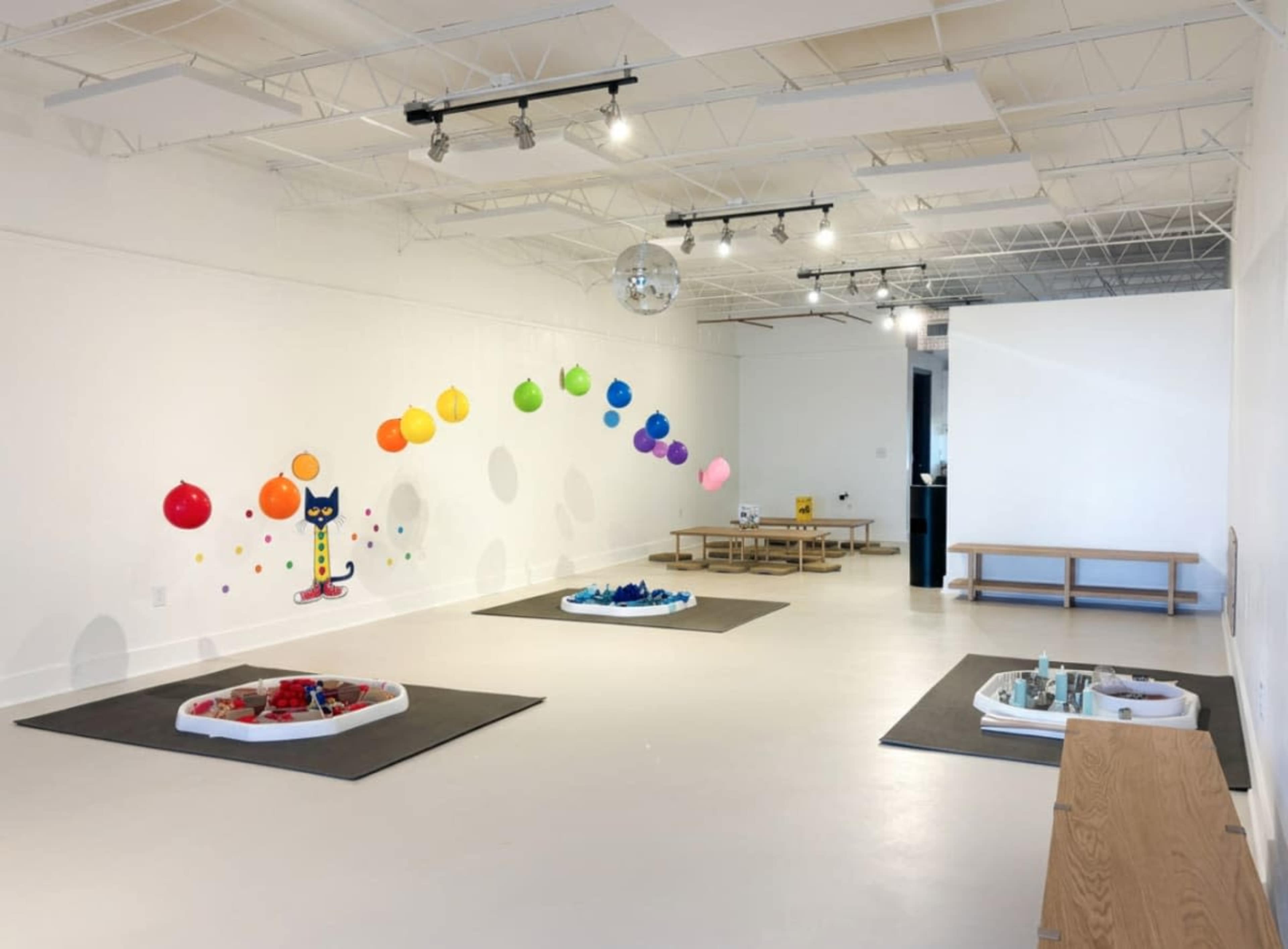 The image shows a brightly lit, empty room featuring a colorful wall installation of balloons, two play areas with toys, and several benches along the walls.