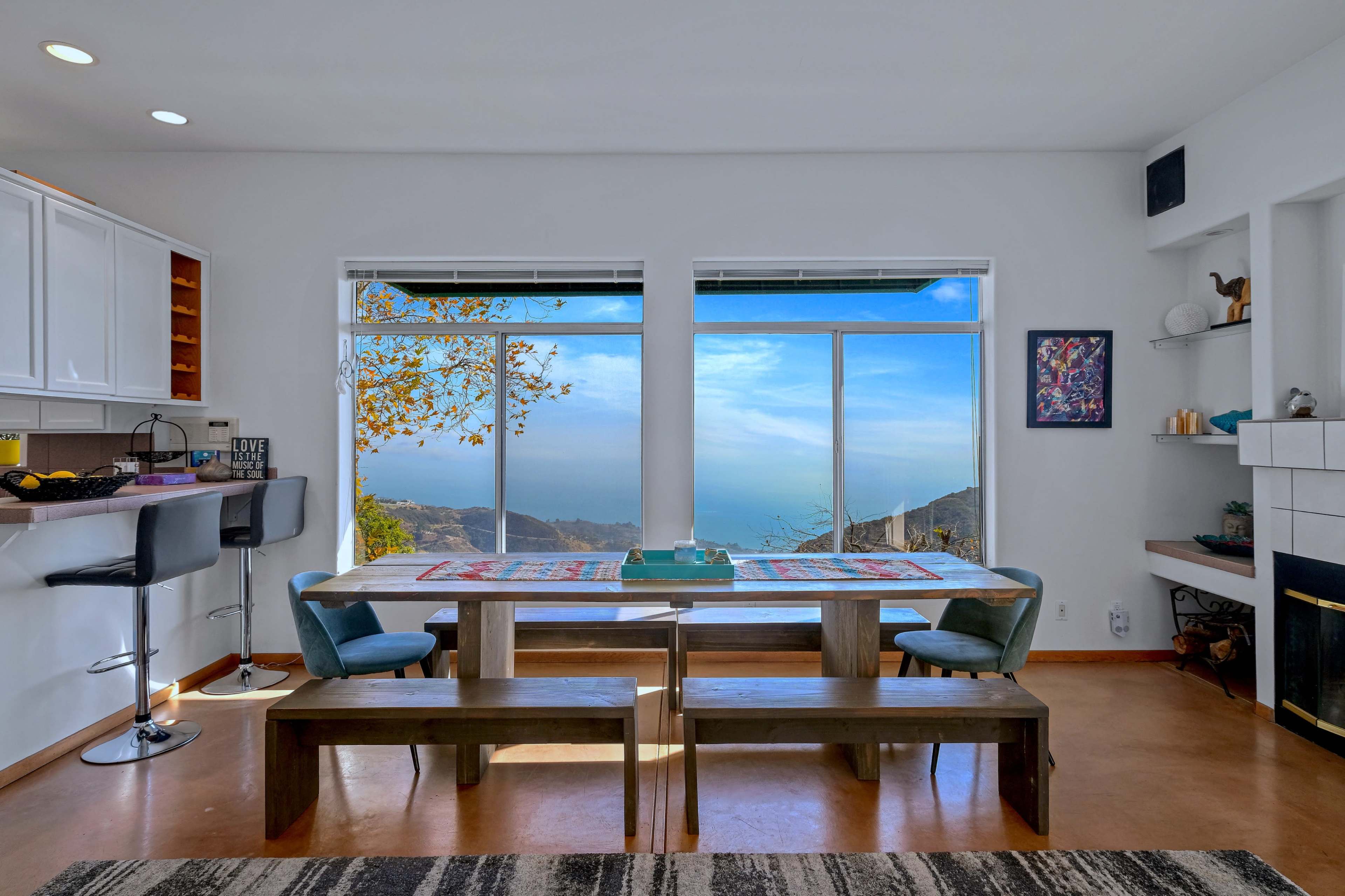The image showcases a modern dining area with a long wooden table, benches, and large windows overlooking a scenic ocean view.