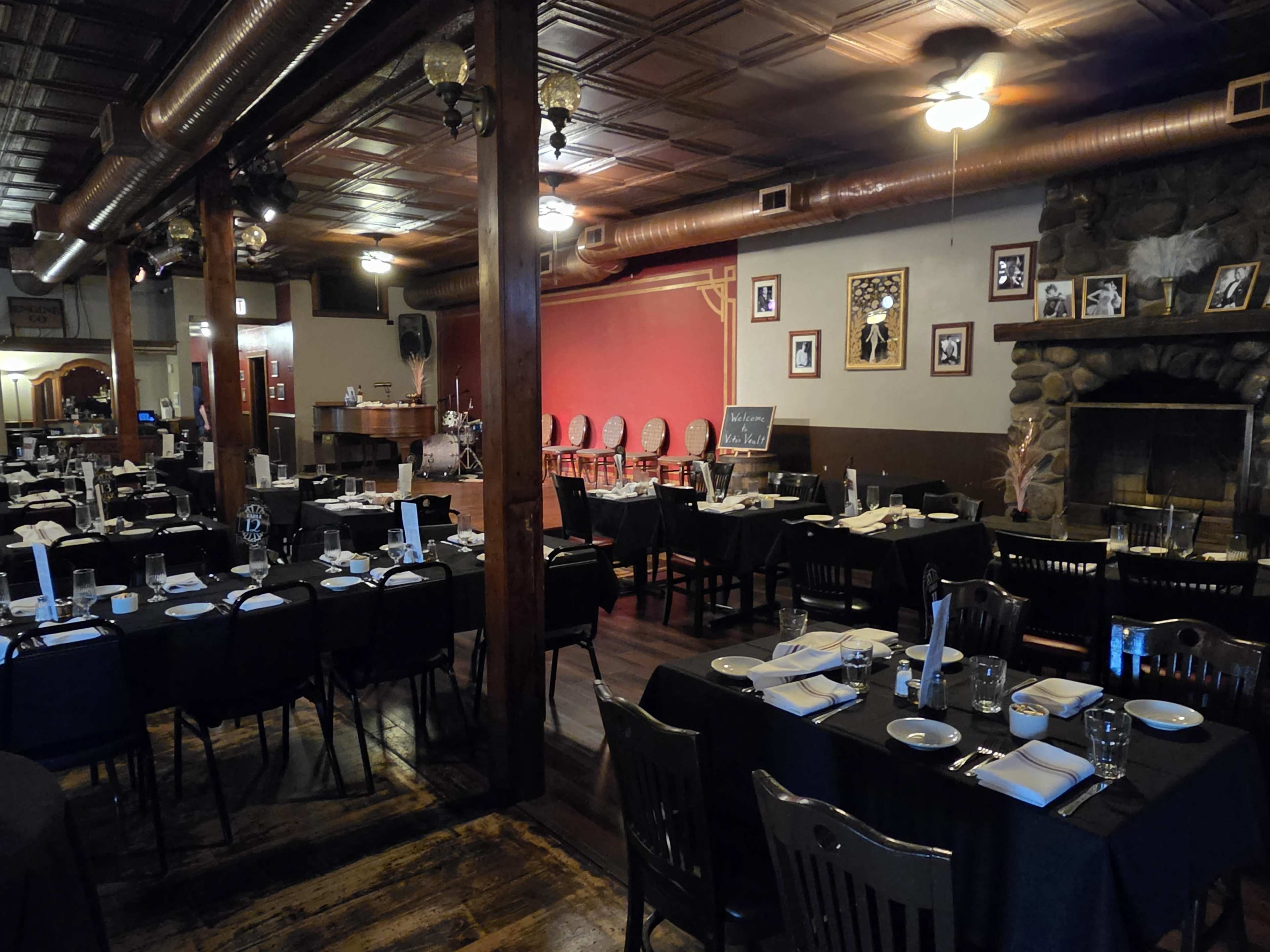 The image shows a restaurant interior with black-covered tables set for dining, a stage area at the end, and a stone fireplace.