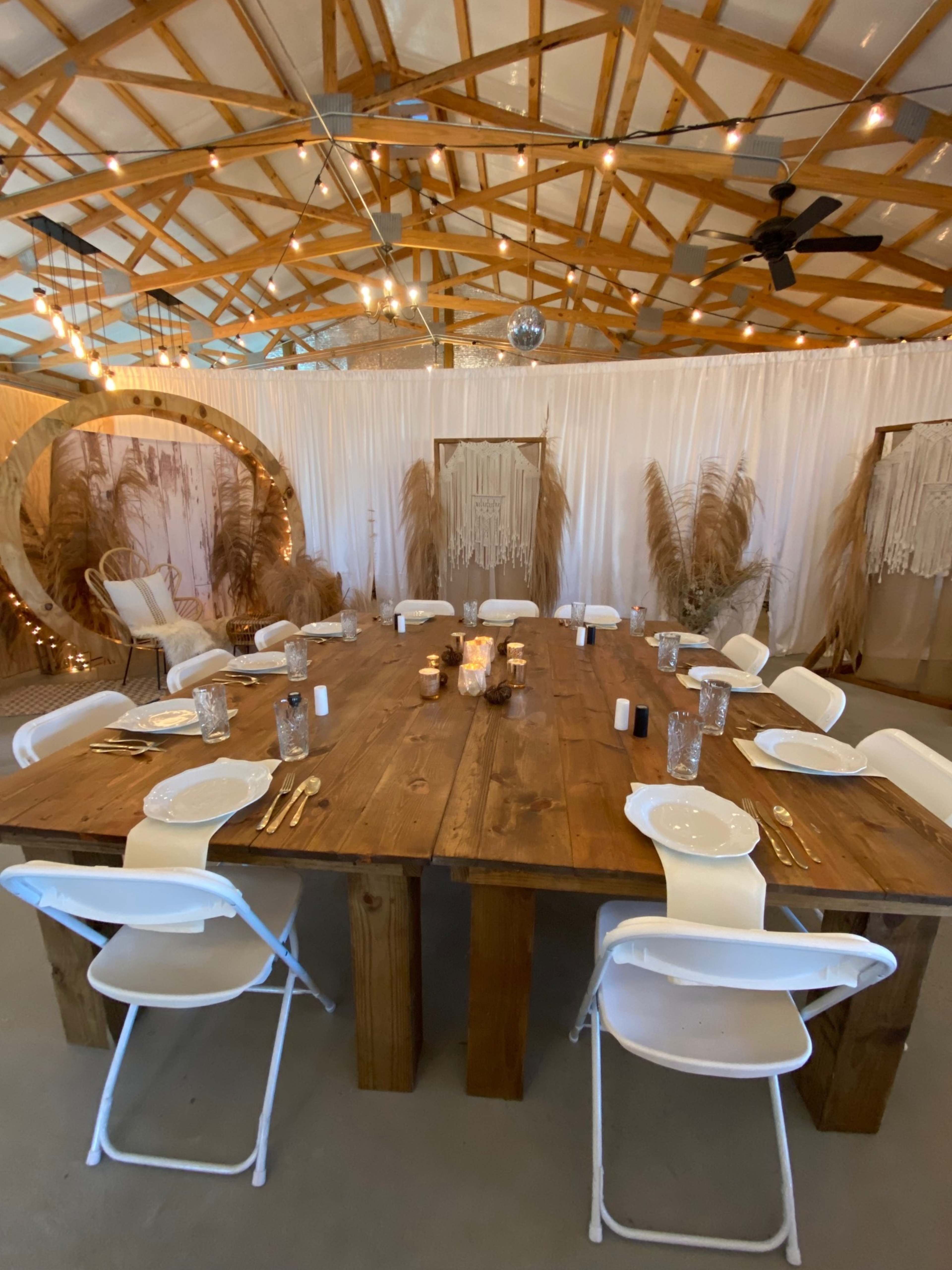 A long wooden table is set with white plates and glasses, surrounded by white folding chairs, in a decorated indoor space featuring string lights and decorative backdrops.
