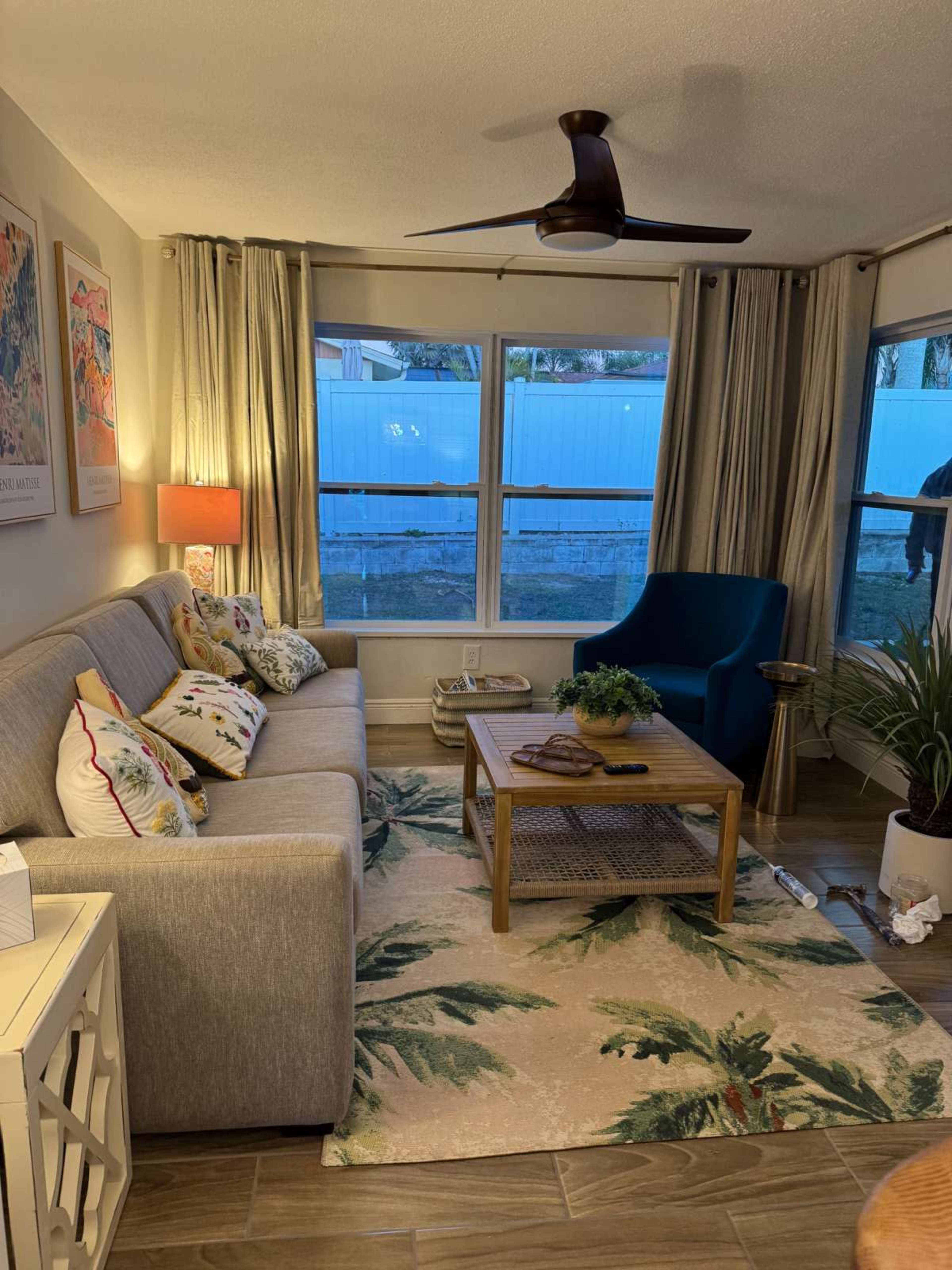 The living room features a gray sofa, a green accent chair, a coffee table with decorative items, and large windows with curtains.