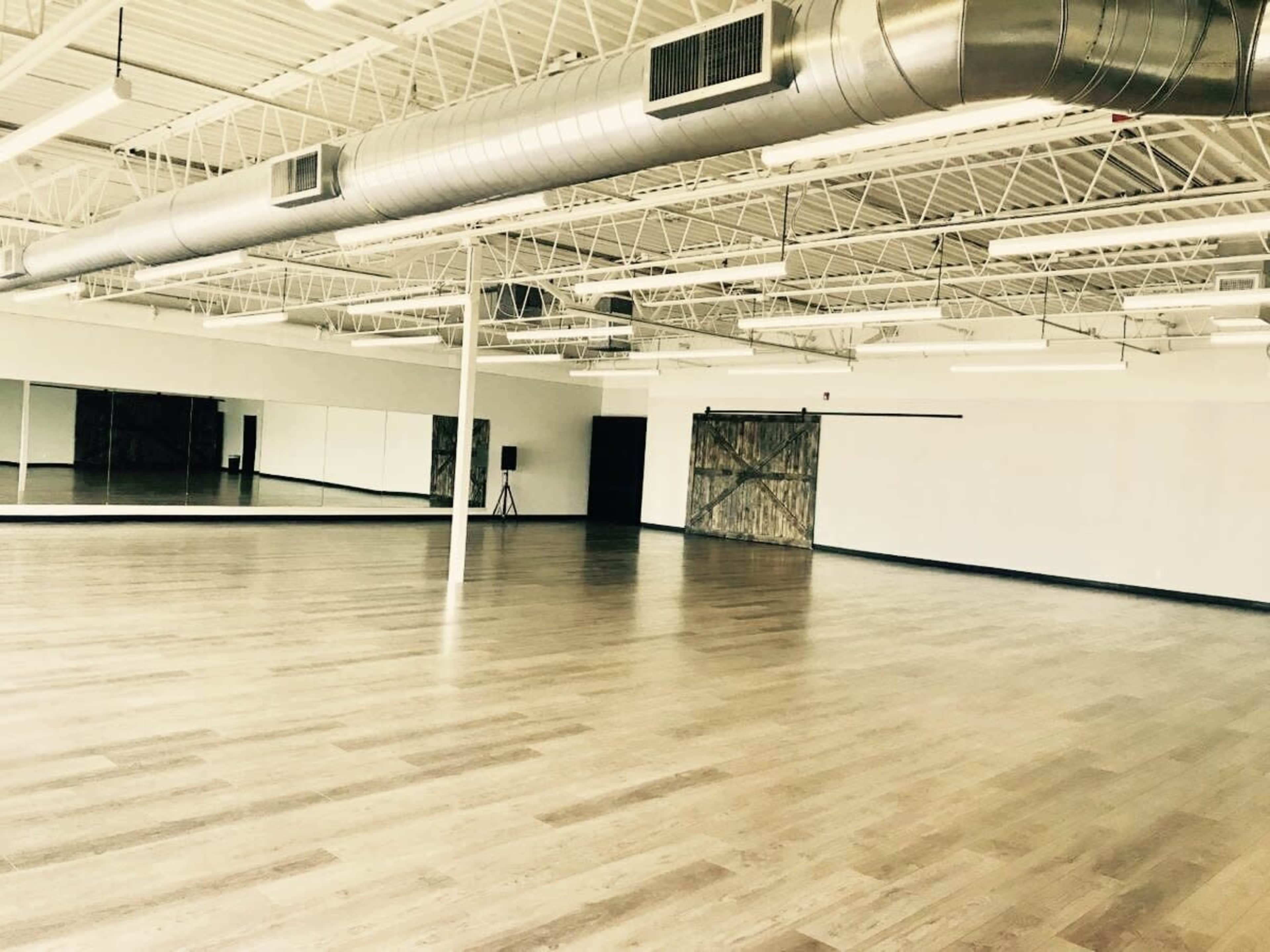 The image shows a spacious dance studio with wooden flooring, large mirrors, and industrial-style ventilation.