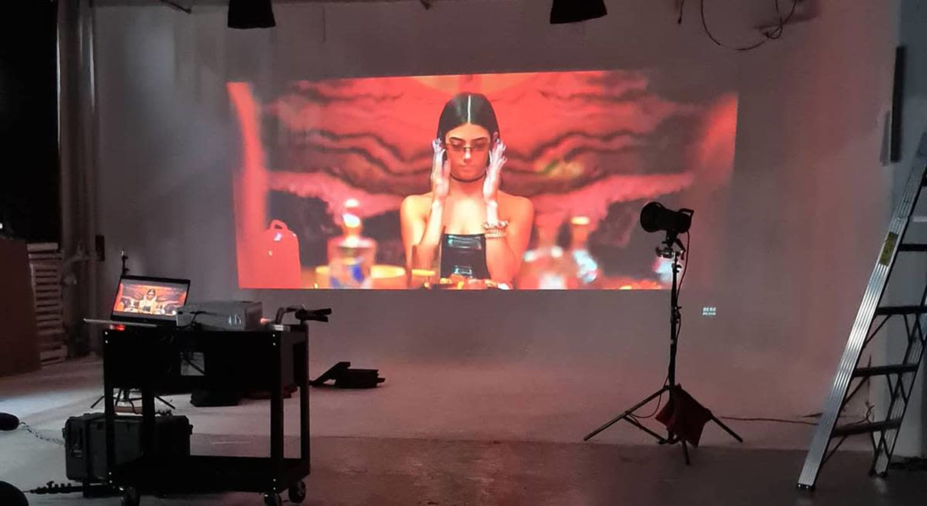 A film or video shoot is taking place in a studio, featuring a projection of a woman in front of a colorful backdrop, with lighting equipment and a production setup visible.