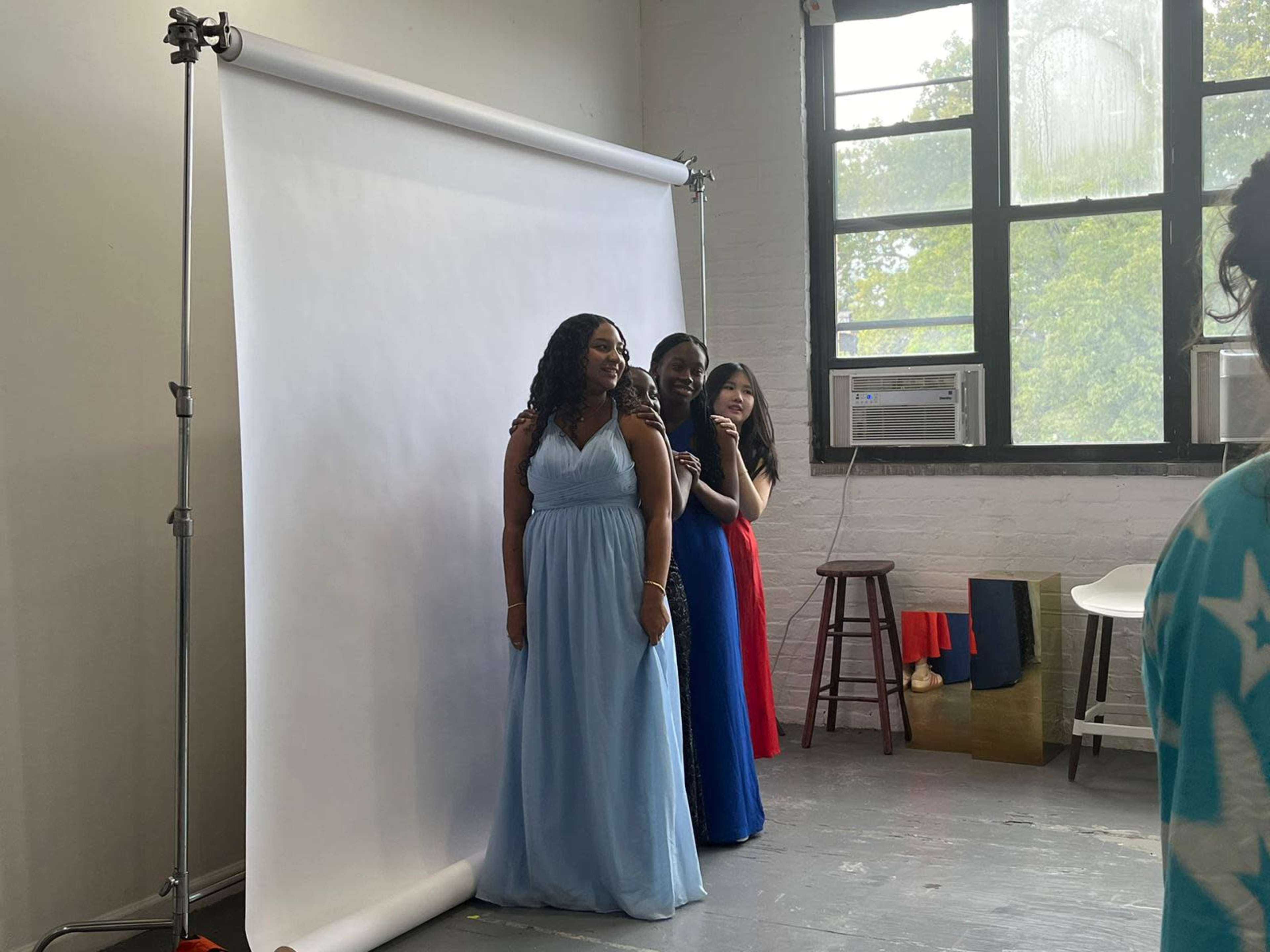 Three women are posing in front of a white backdrop, wearing long dresses in blue and red.