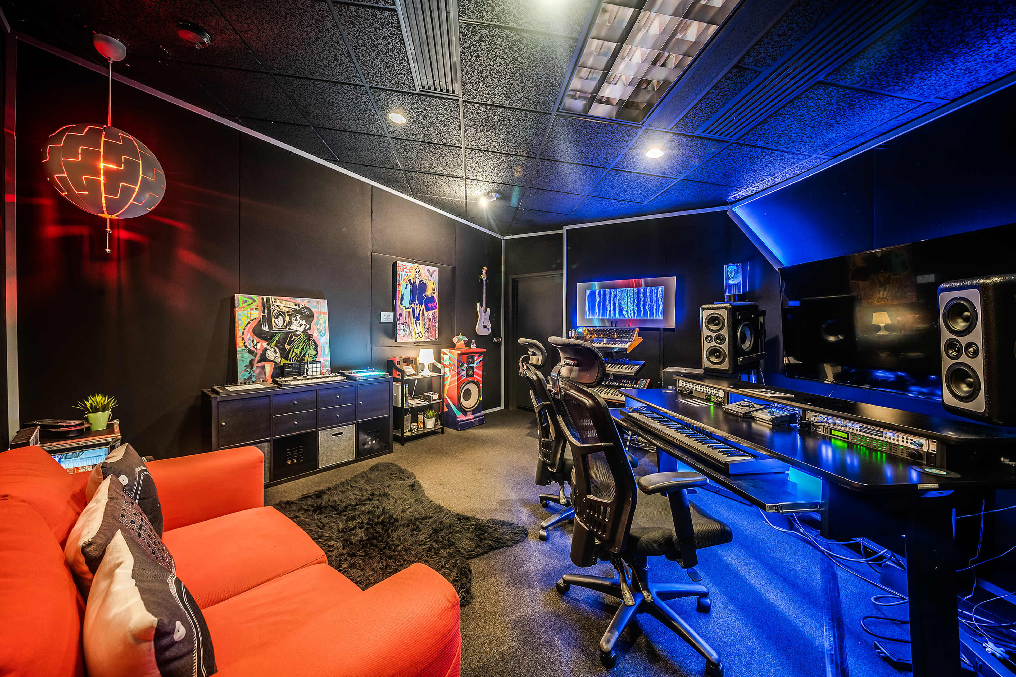 A modern music studio featuring a black wall, a computer desk with audio equipment, a red sofa, and vibrant decorative elements.