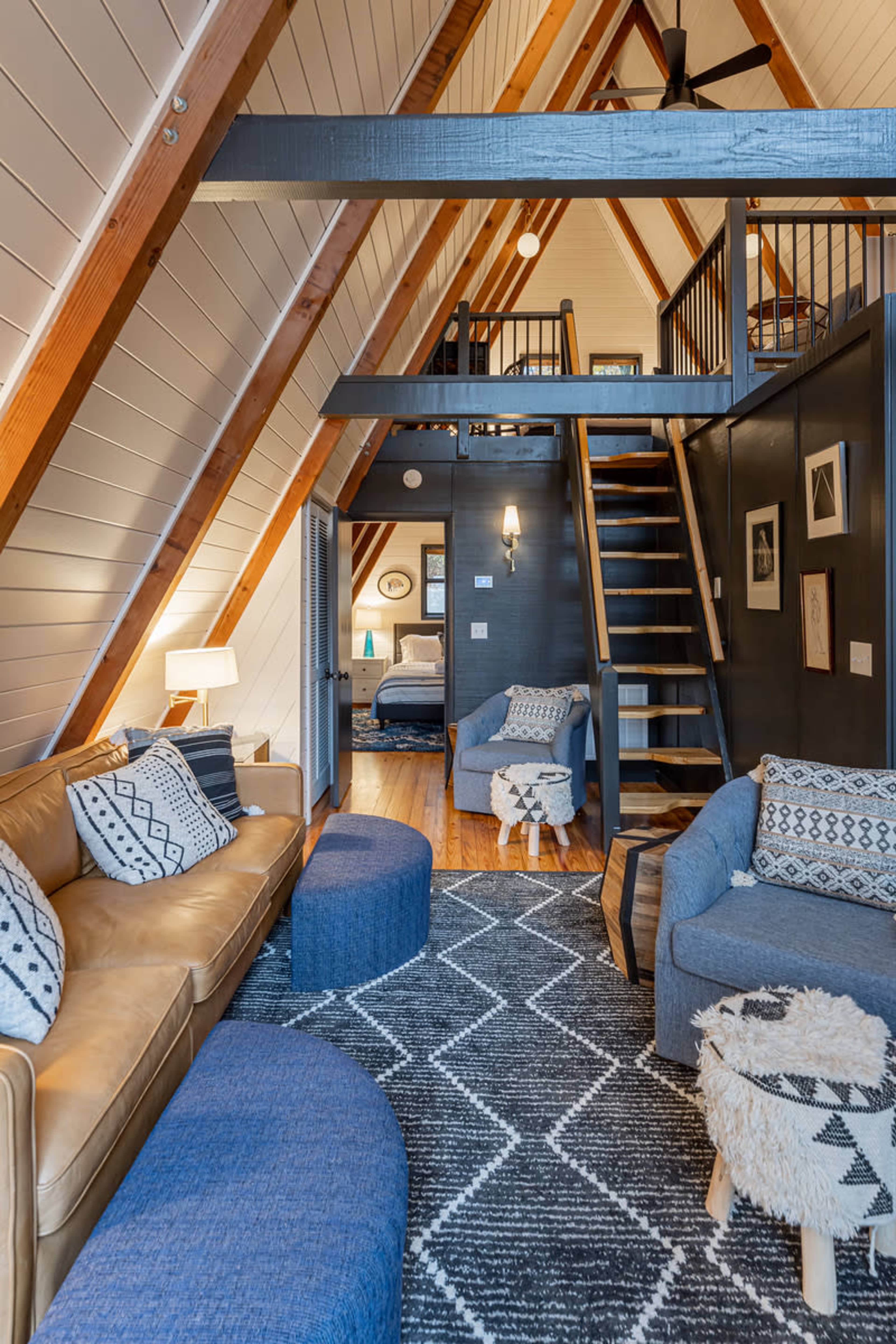 The image shows a cozy, contemporary living area in a chalet-style home, featuring a mix of leather and fabric seating, a patterned rug, and an open staircase leading to an upper level.