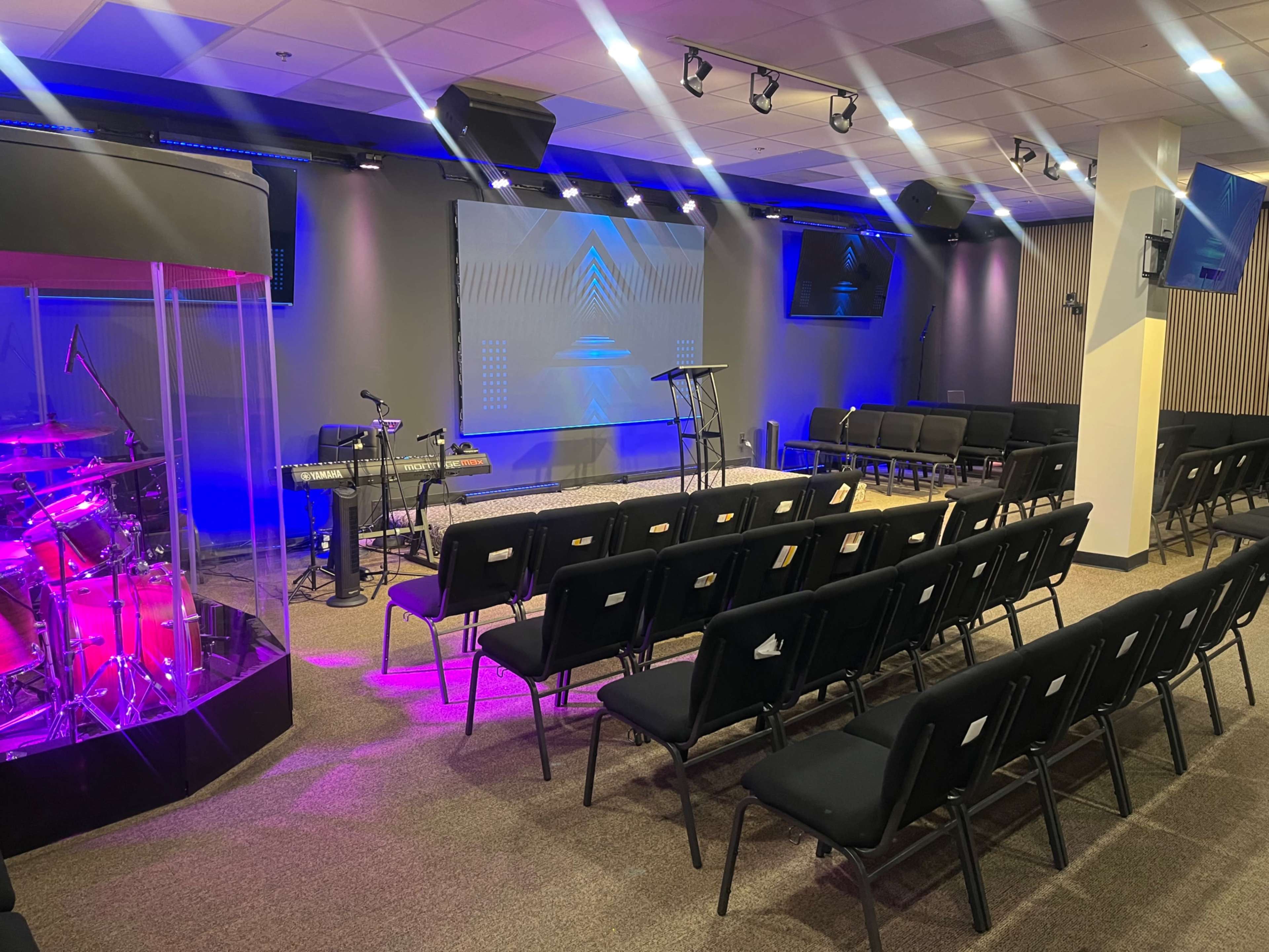 The image shows a modern worship space with rows of black chairs facing a central stage, flanked by musical instruments and a large screen in the background.