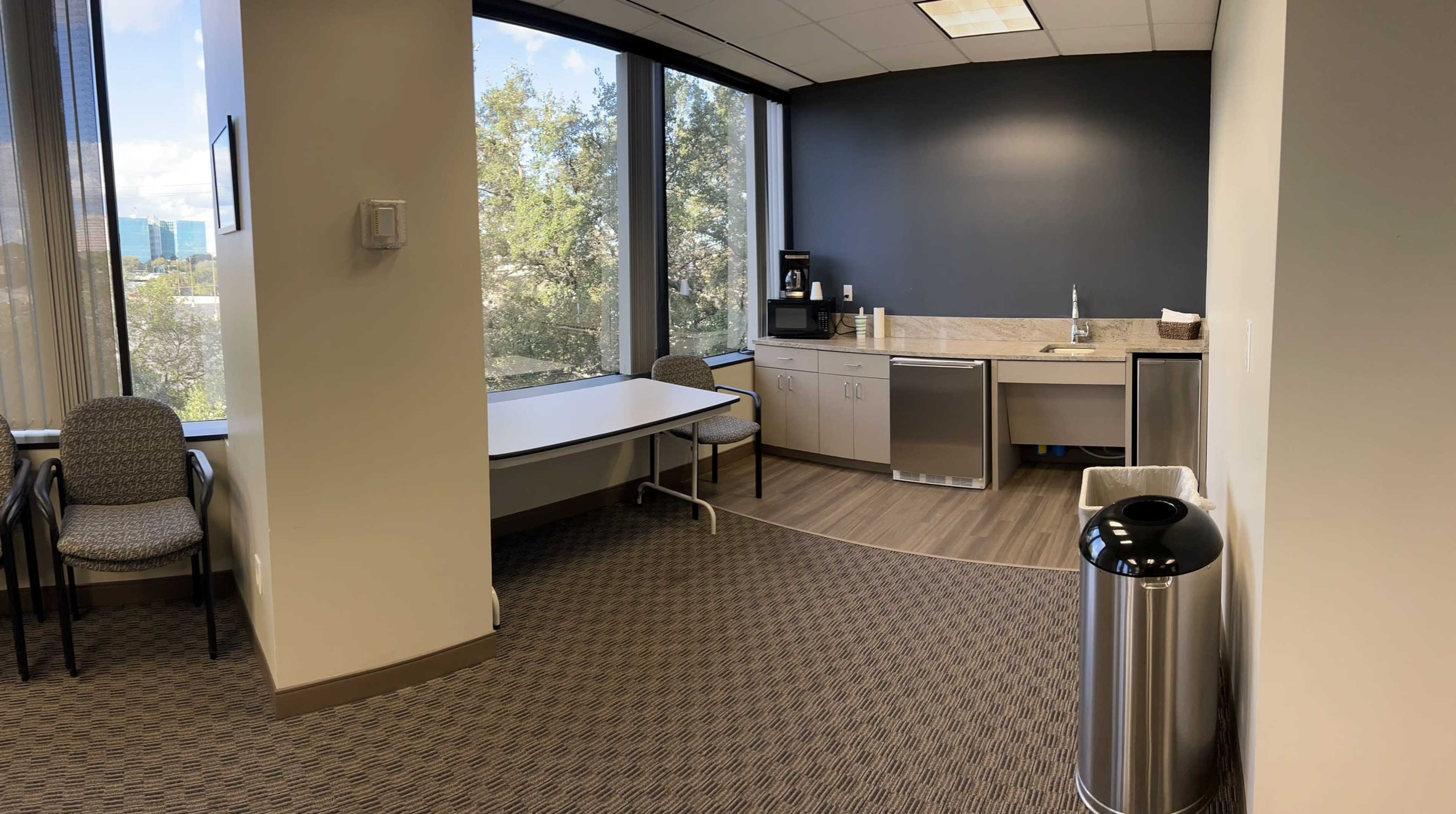An office break area with a kitchenette, including a sink, refrigerator, and counter, alongside a table and chairs near large windows.