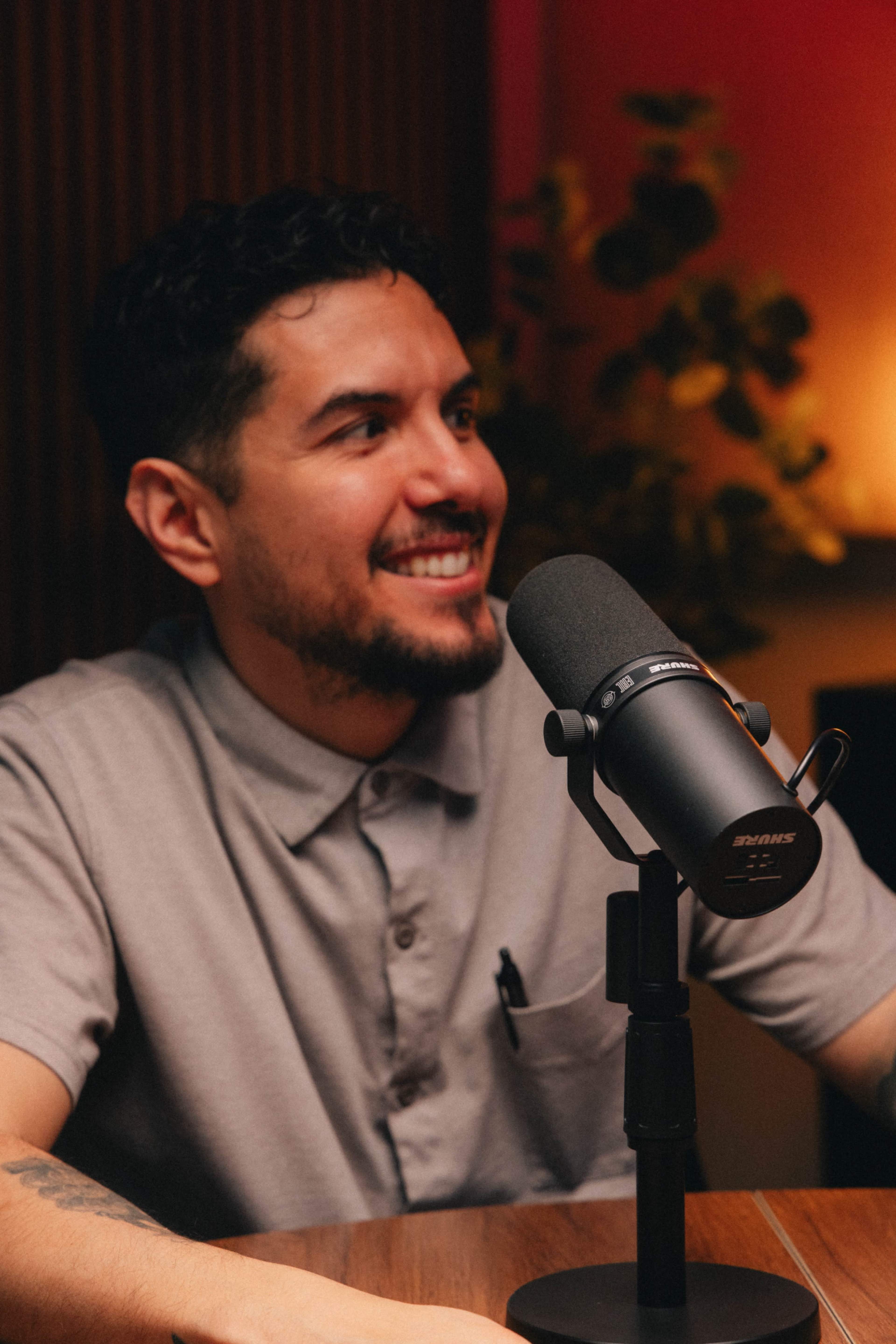 A man with dark hair and a light gray shirt smiles while sitting at a table with a microphone in front of him.
